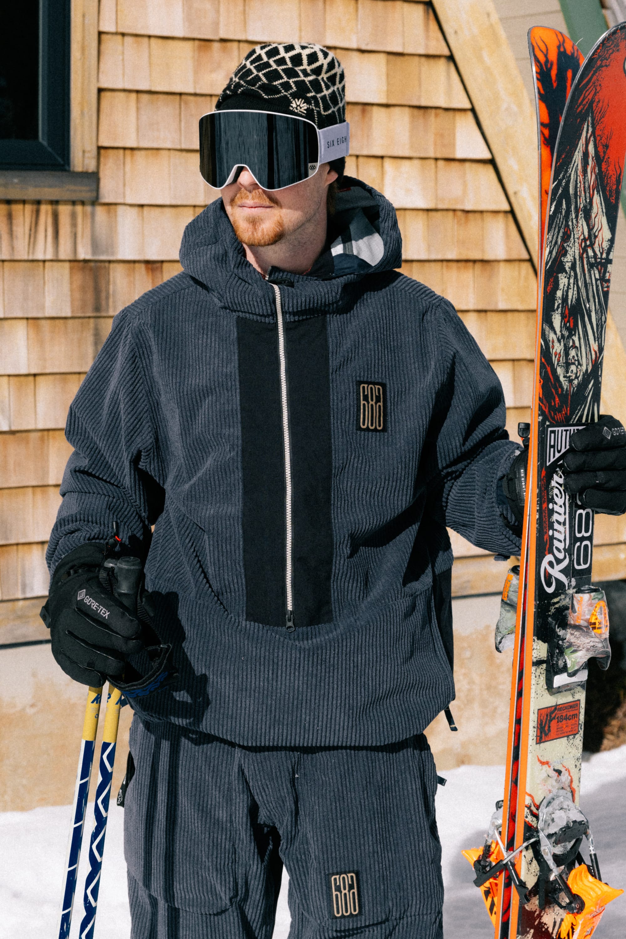 Wearing the 686 Men’s Ghost™ 2.5L Anorak by 686, a person stands outside on snow holding skis and poles, with a wooden building in the background.
