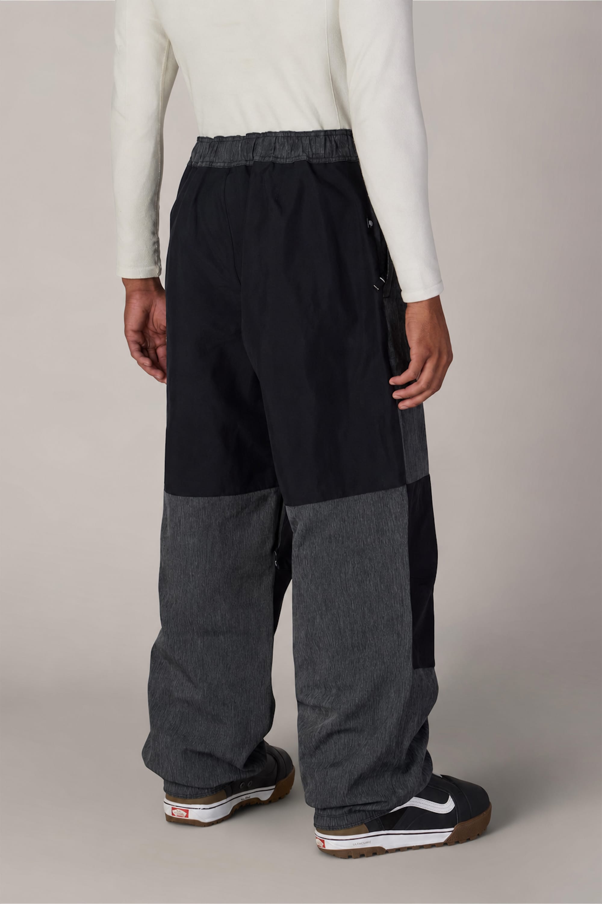 A person wearing loose-fitting 686 Ghost™ 2.5L Pant by 686 in black and gray, a white long-sleeve shirt, and black Nike sneakers with white soles is standing against a plain background, viewed from behind.