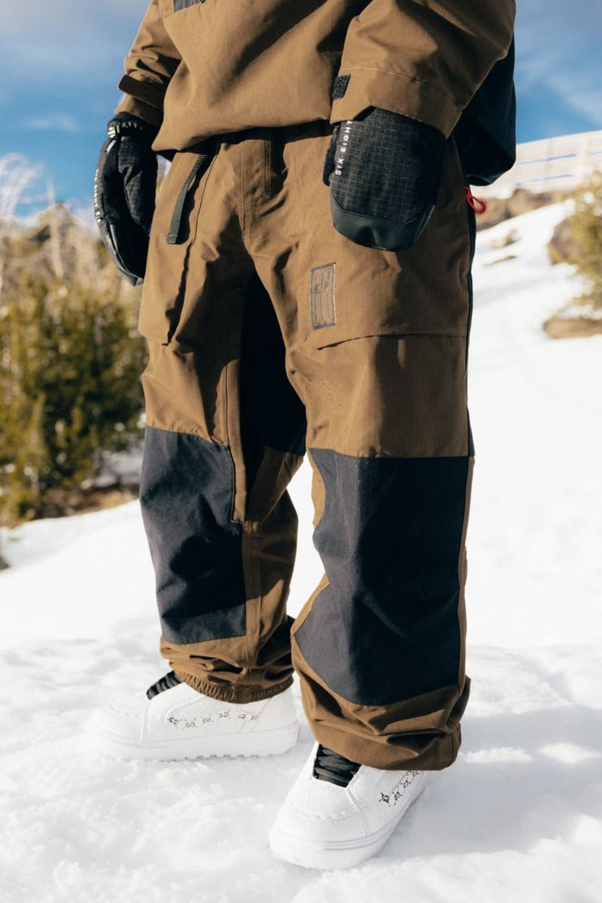 A person wearing the 686 Ghost™ 2.5L Pant by 686, along with black gloves and white sneakers, stands on snow with green bushes and a wooden fence in the background.