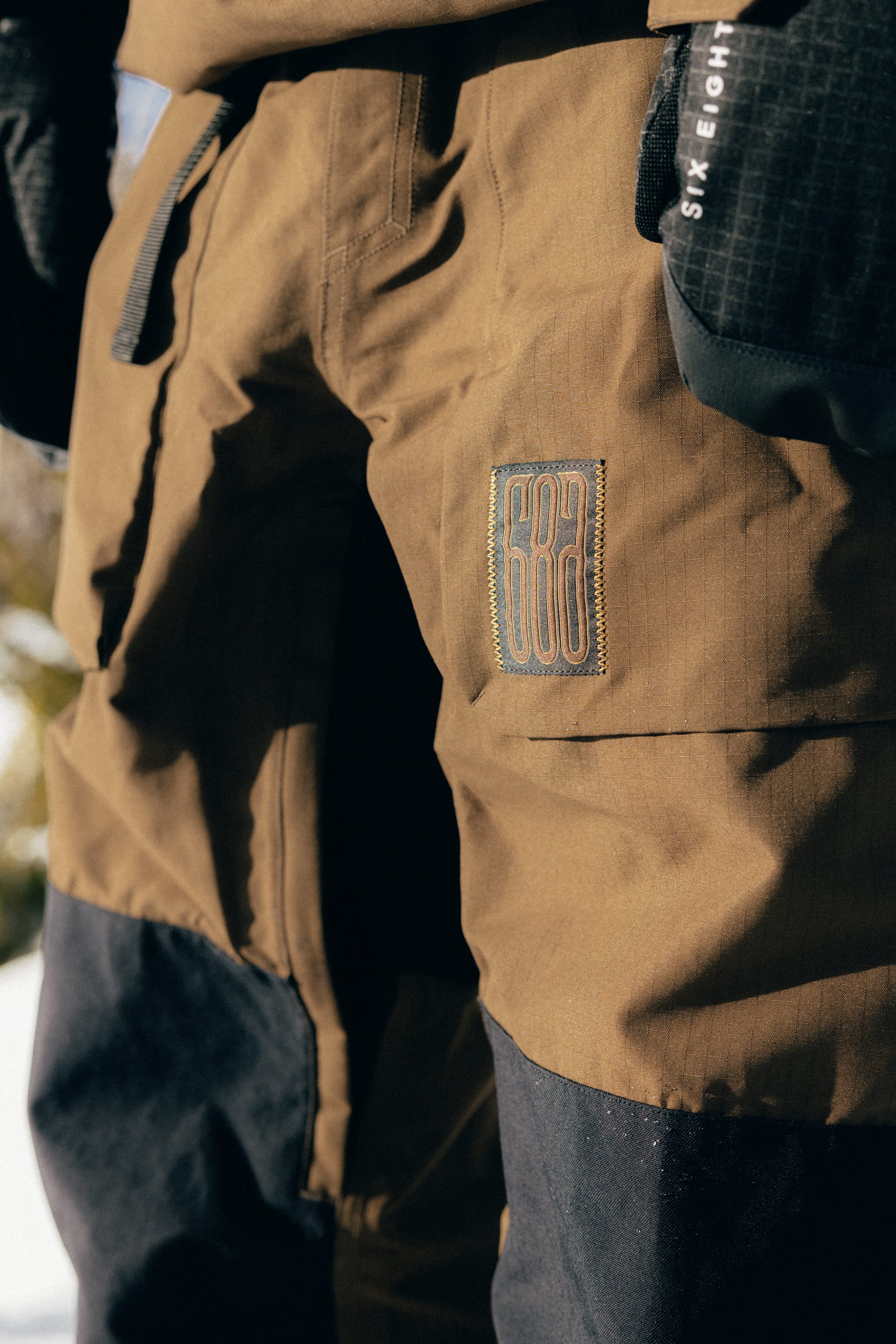 A close-up of a person wearing the 686 Ghost™ 2.5L Pant, featuring brown and black color-blocked panels with a 686 logo patch, paired with black gloves and standing outdoors in sunlight.