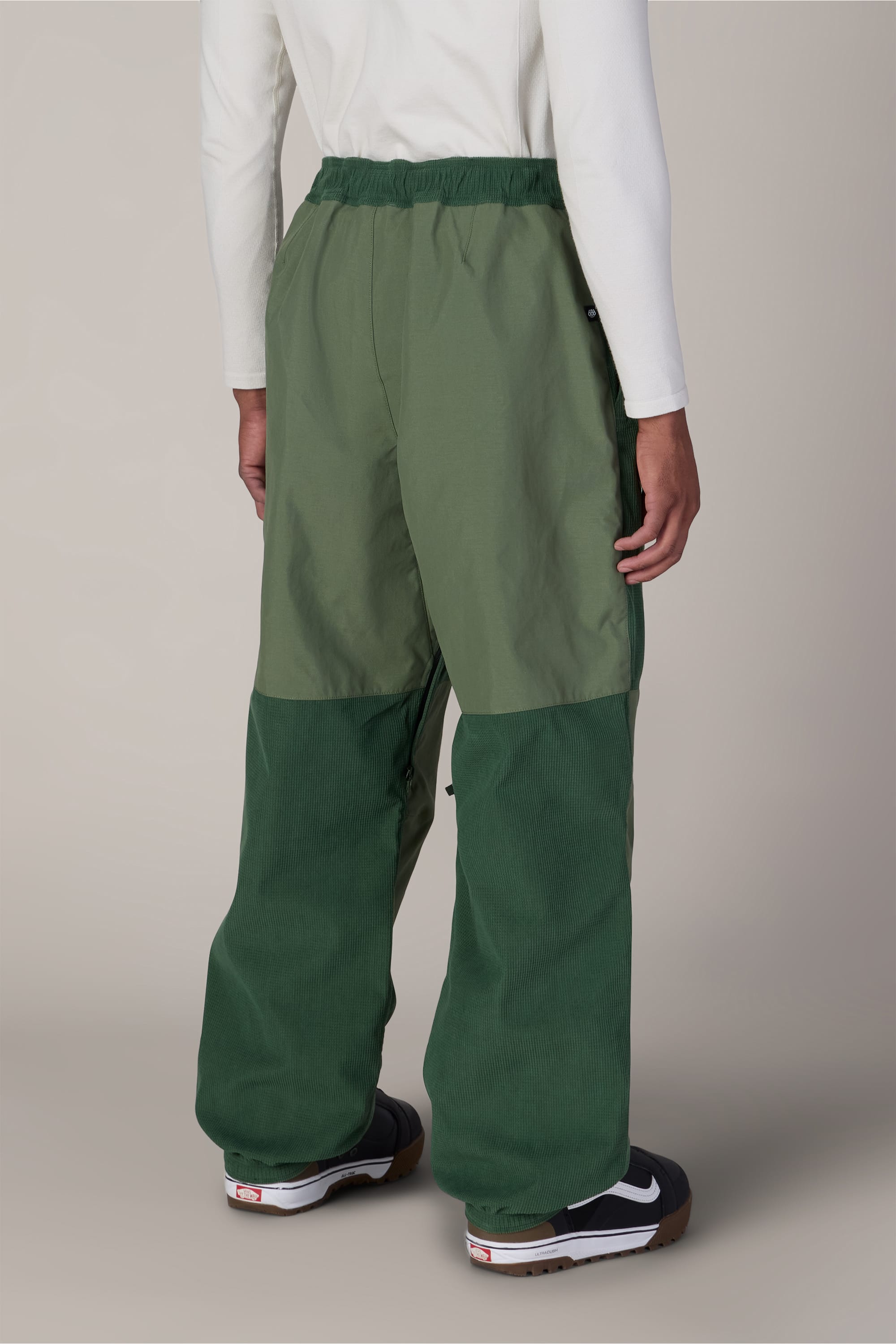 A person wearing the 686 Ghost™ 2.5L Pant by 686 in a two-tone green color, paired with a long-sleeve white shirt and black Nike sneakers, shown from the back against a neutral background.