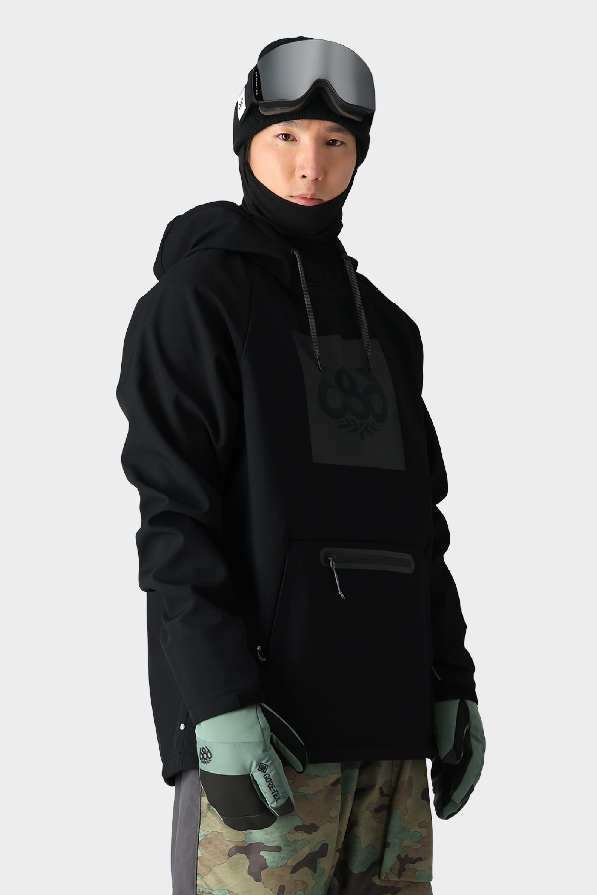 A person wearing the 686 Men’s Waterproof Hoody by 686 faces forward against a plain light background, styled with ski goggles, green gloves, and camouflage pants.