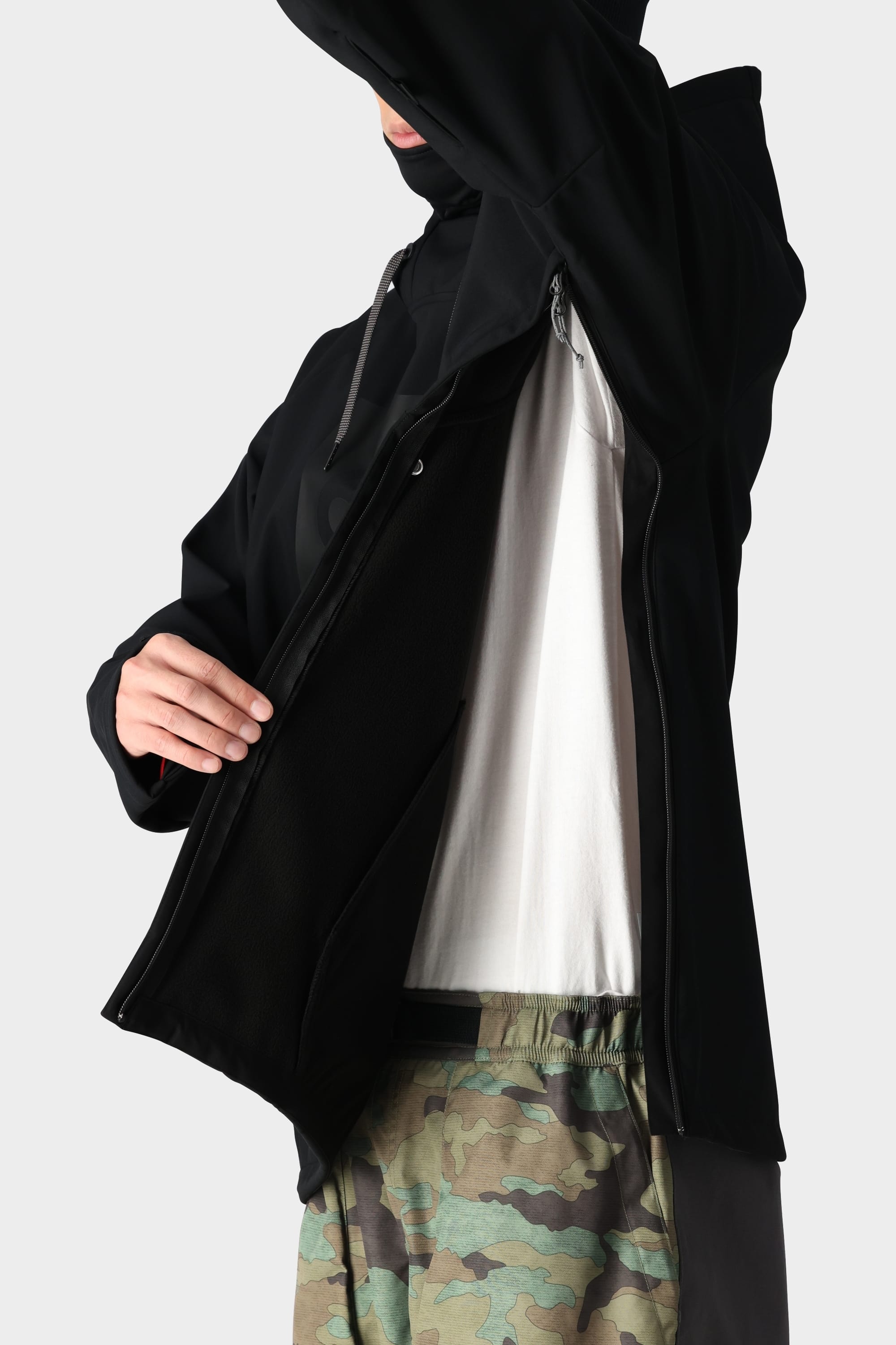 A person wearing the 686 Men’s Waterproof Hoody by 686, layered over a white shirt and paired with camouflage pants, opens the jacket to display its inner lining. Their face is partially out of the frame.