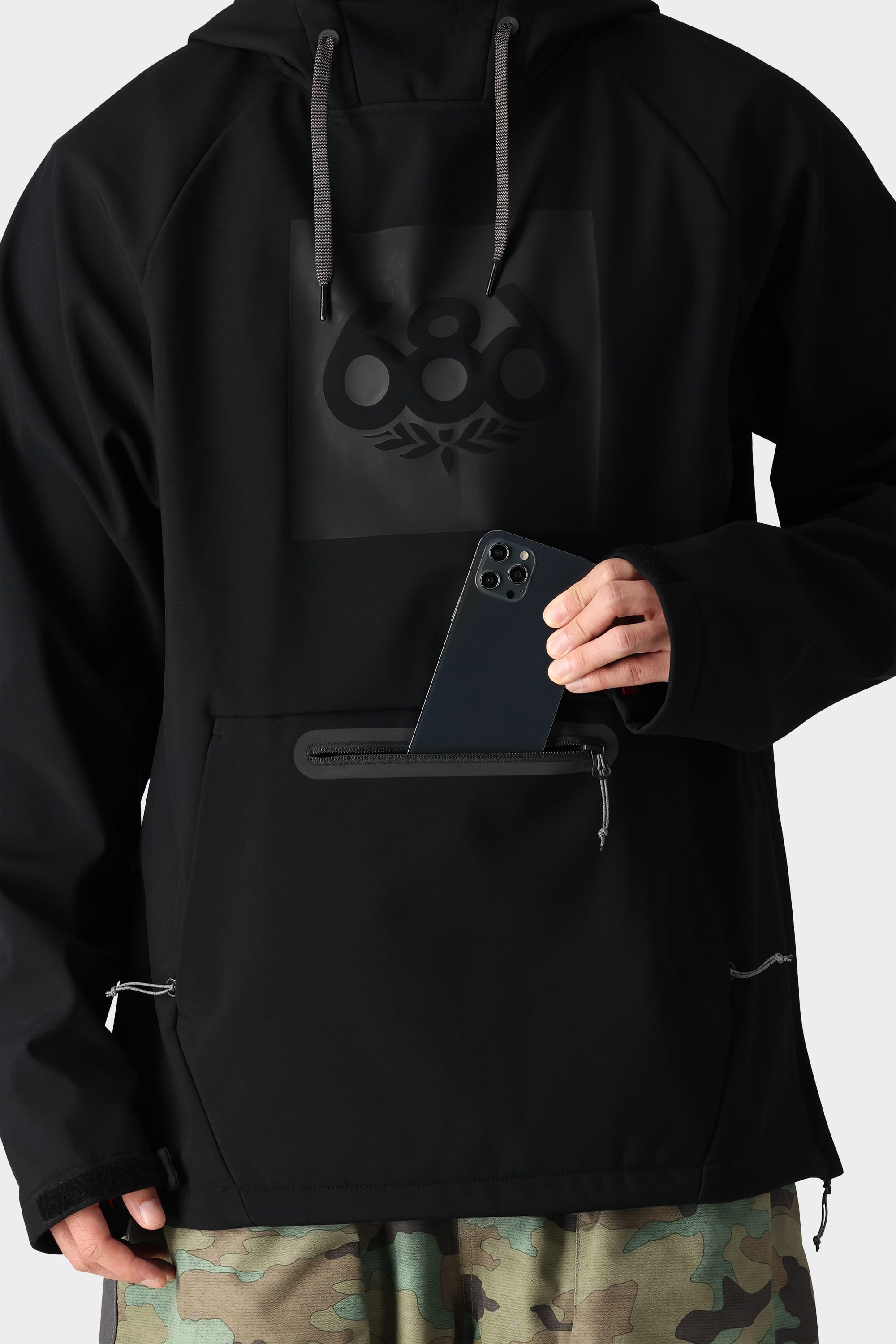 A person wearing the 686 Men’s Waterproof Hoody in black, featuring a logo, and camo pants places a black smartphone into the front zippered pocket of the hoodie. Only their torso and hand are visible against a plain background.