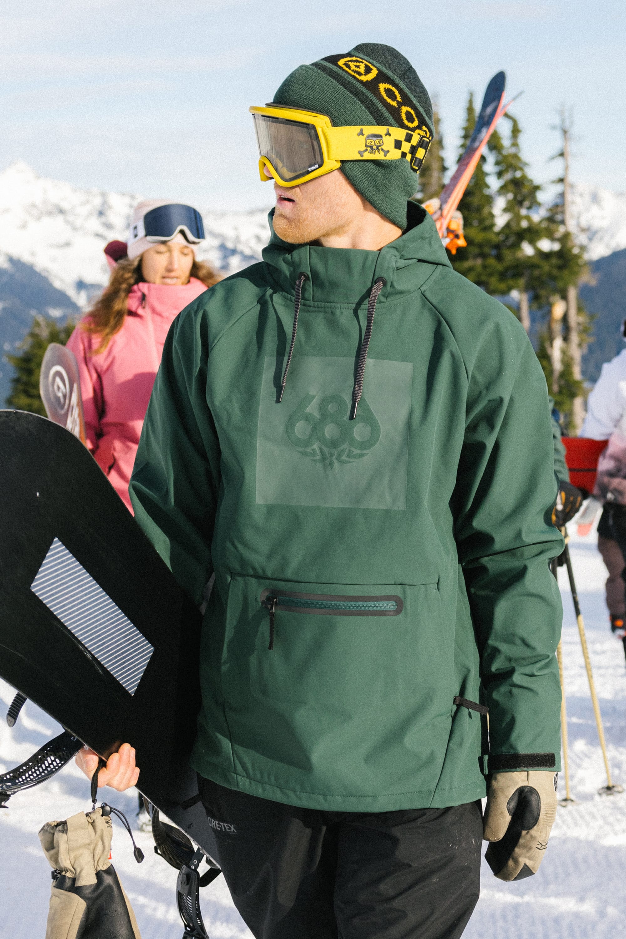 Wearing a 686 Men’s Waterproof Hoody, a person in yellow goggles holds a snowboard on a snowy mountain, with other skiers and snowboarders in the background.