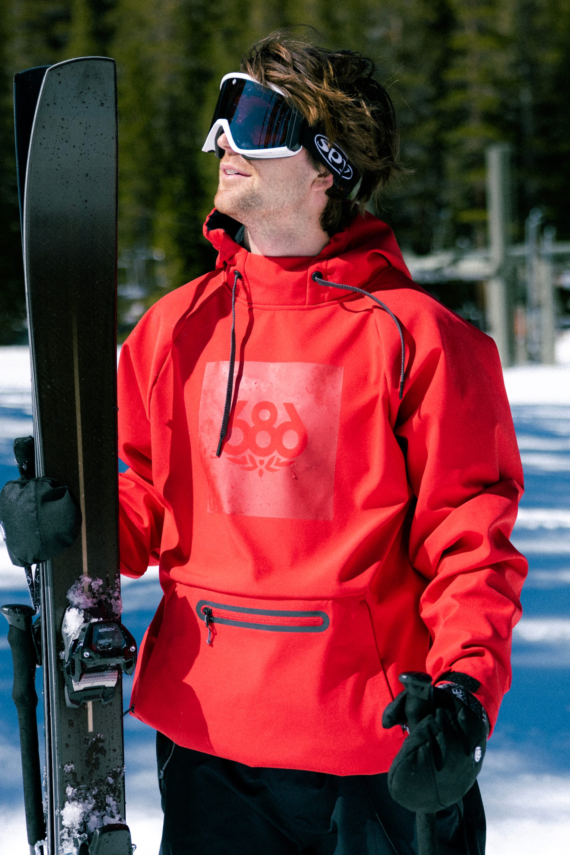 Wearing the 686 Men’s Waterproof Hoody, a person stands in the snow with skis, looking upward. Trees and ski equipment from 686 are visible in the background.