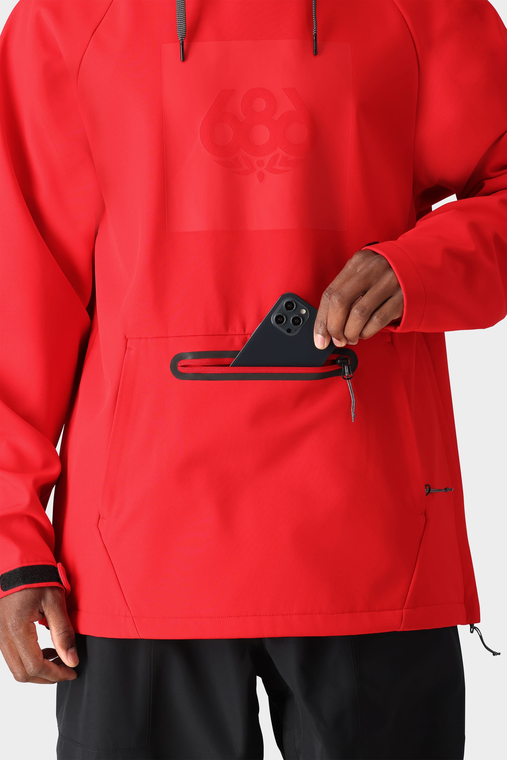 A person wearing the 686 Men’s Waterproof Hoody in bright red uses one hand to put a black smartphone into the horizontal zippered front pocket. Only the torso and hand are visible.