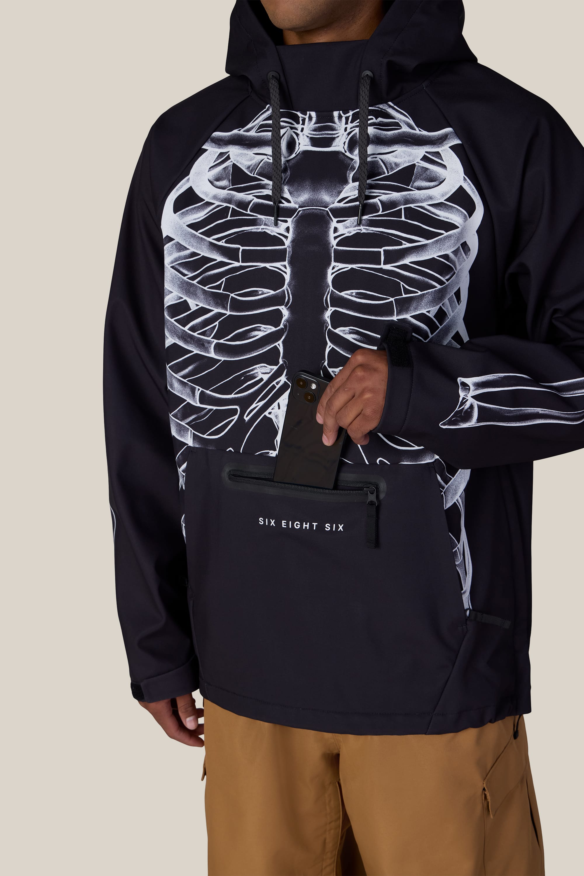A person wearing the 686 Men’s Waterproof Hoody in black with a white ribcage design and tan pants, holding a phone while standing with one hand in a front pocket marked with the 686 logo.