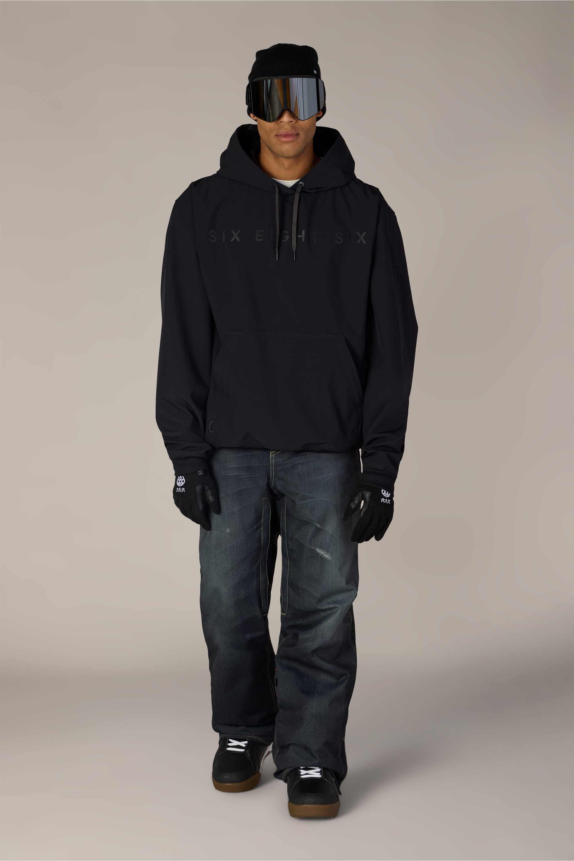 A person is wearing the 686 Men’s Waterproof Team Hoody by 686, along with a black beanie, reflective ski goggles, black gloves, dark jeans, and black sneakers, standing against a plain light background.