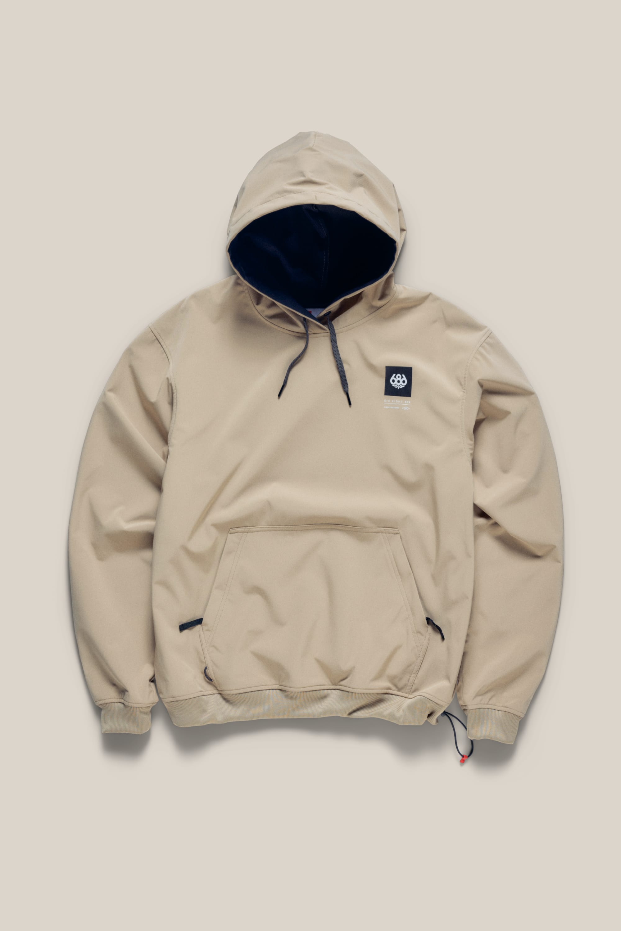 The 686 Men’s Waterproof Team Hoody by 686 is shown laid flat on a plain light background, featuring a beige colorway with black drawstrings, a front pocket, and a small black logo patch on the left chest.