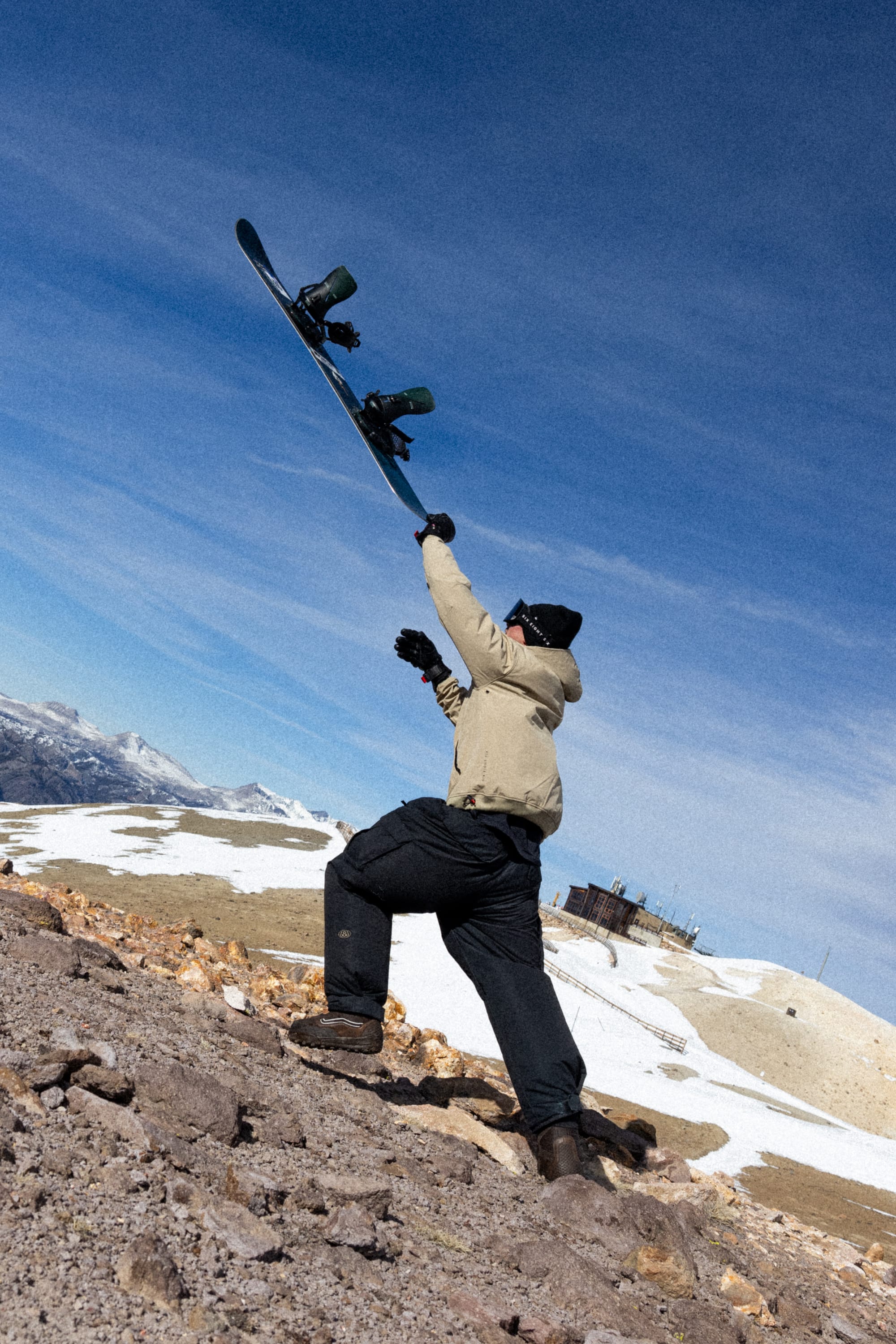 Wearing the 686 Men’s Waterproof Team Hoody, a person in winter gear lifts a snowboard triumphantly while standing on a rocky, snow-patched hillside under a blue sky with distant mountains.