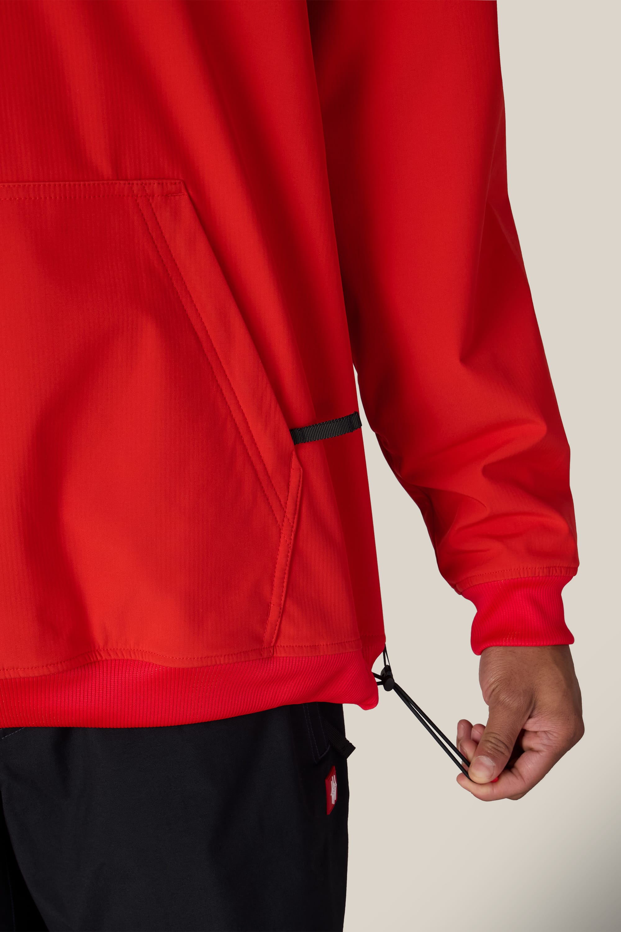 Someone wearing the 686 Men’s Waterproof Team Hoody adjusts the hem drawstring near the side pocket, with only their hand and part of their body visible against a plain background.