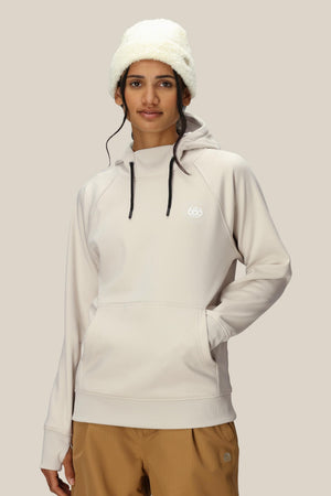 A woman in a cream-colored 686 Womens Bonded Fleece Pullover Hoody with a chest logo, tan pants, and a fluffy white hat stands against a beige background, looking at the camera with a neutral expression.