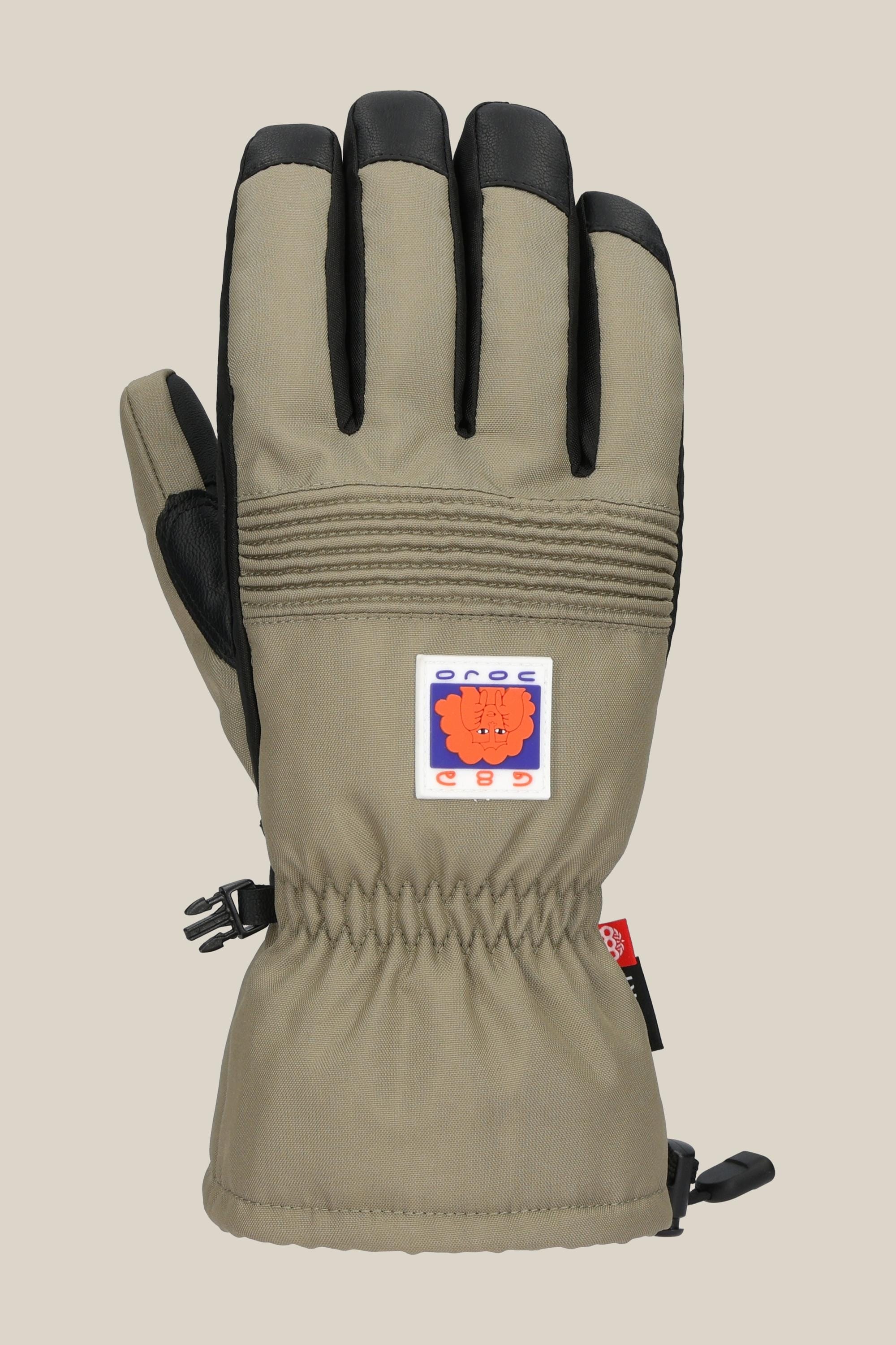 The 686 Lander Glove by 686 is a beige winter glove with black fingertips and palm, a colorful orange bear patch on the back, light insulation for warmth, gathered wrist elastic, and a black side clip.