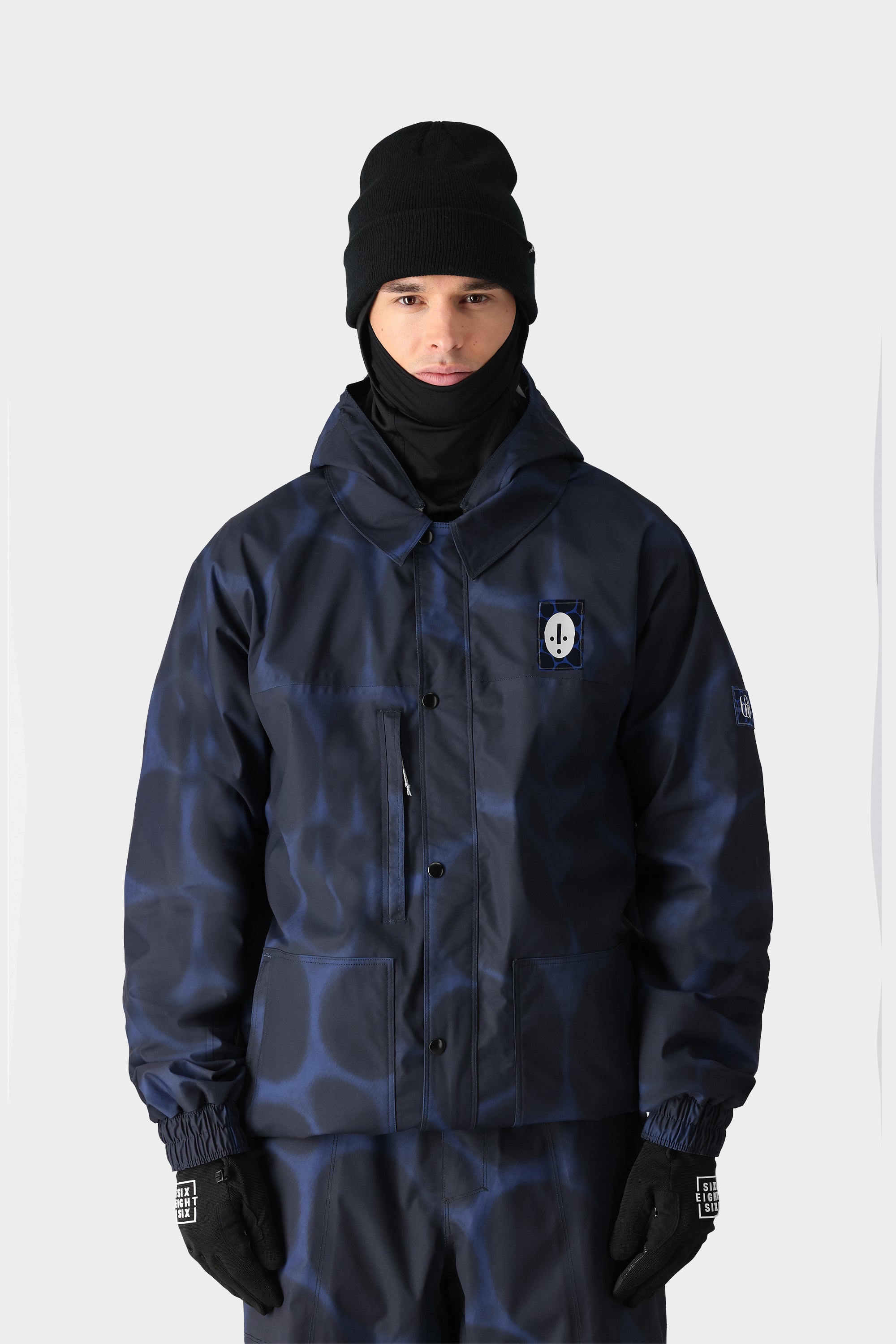 A person wearing the 686 Dojo Jacket by 686, featuring a dark blue pattern and technical details, paired with black gloves, a black beanie, and a black neck gaiter stands against a plain light background.