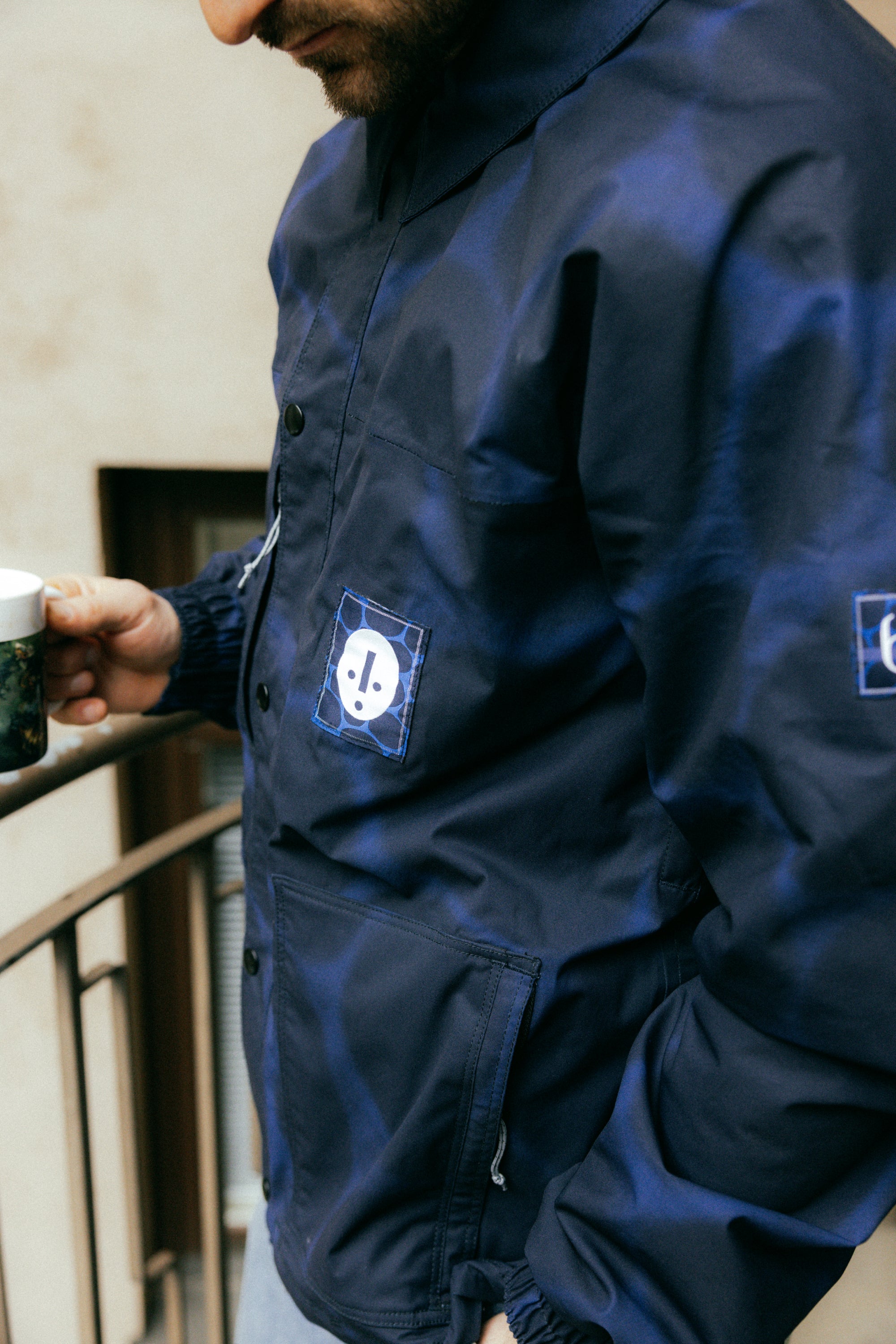 Someone wearing the 686 Dojo Jacket by 686, a dark blue jacket with a white patch design inspired by Forest Baileys technical features, holds a mug on a balcony. The persons face is partly out of frame against an out-of-focus building backdrop.