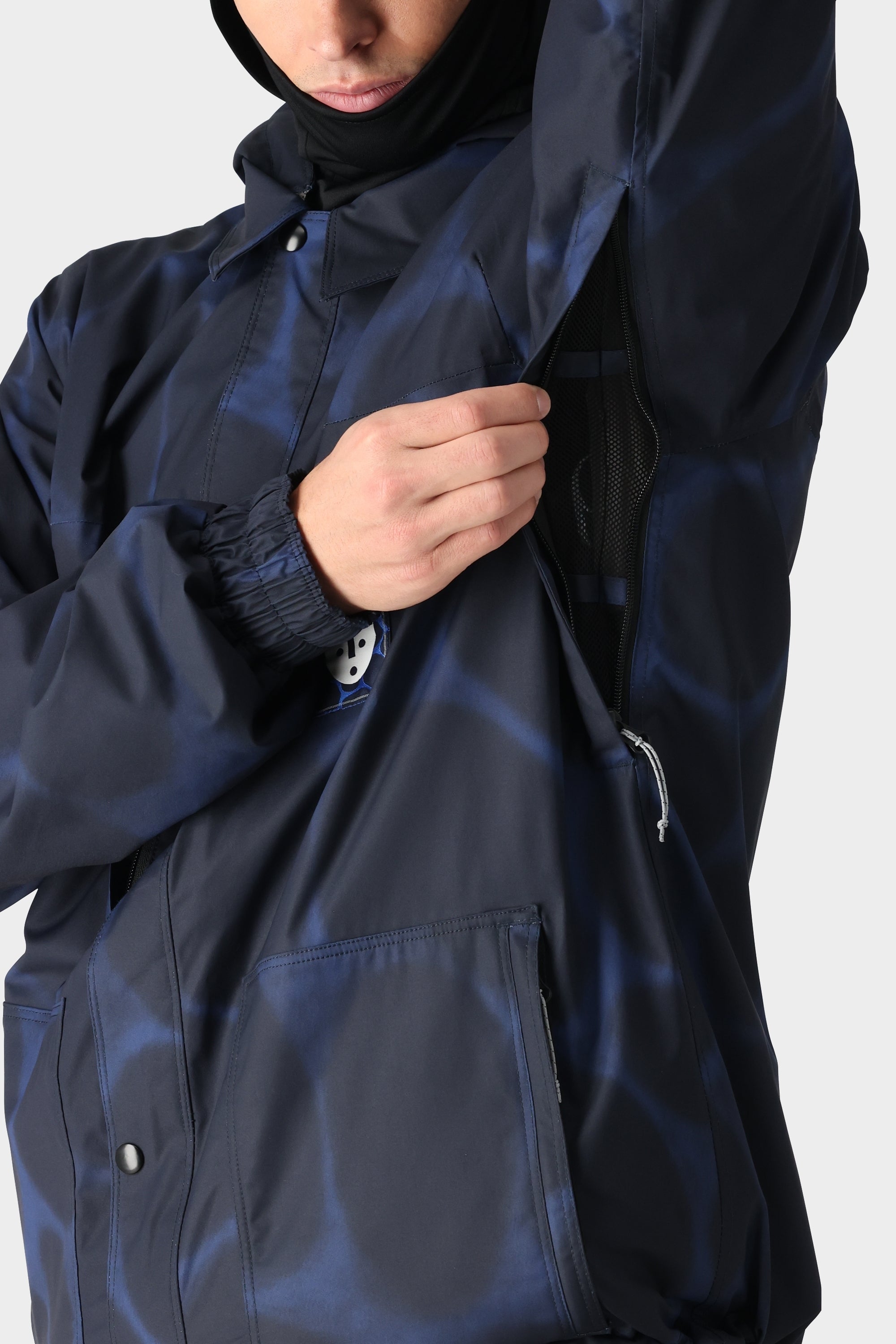 A person wearing the 686 Dojo Jacket from 686 unzips an underarm vent, revealing black mesh lining. Only the lower face is visible, drawing attention to this technical feature of the dark blue patterned jacket.