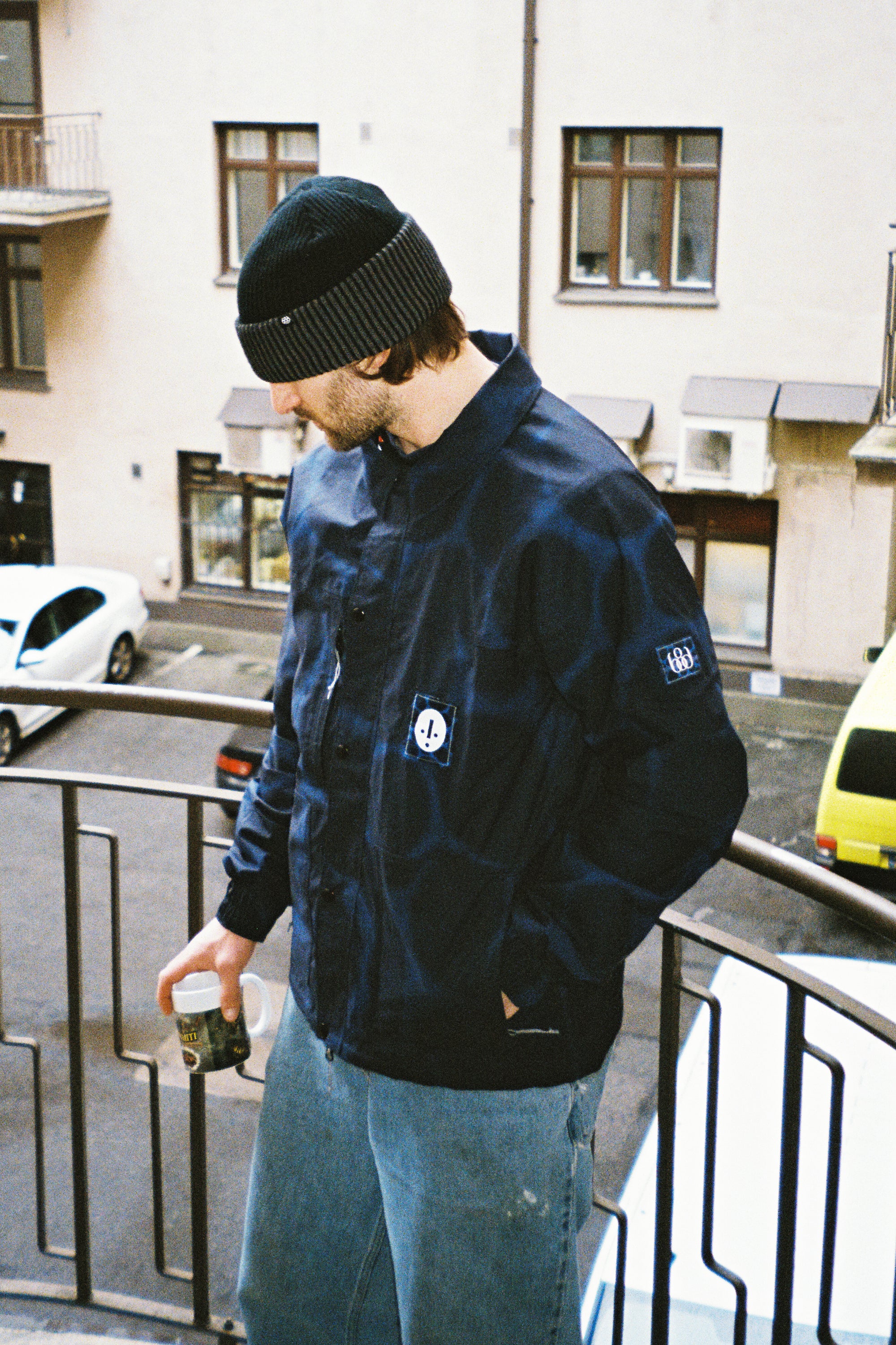 Wearing a black beanie, 686 Dojo Jacket by 686, and blue jeans, a person stands on a balcony holding a drink can. Parked cars and a beige building can be seen in the background.