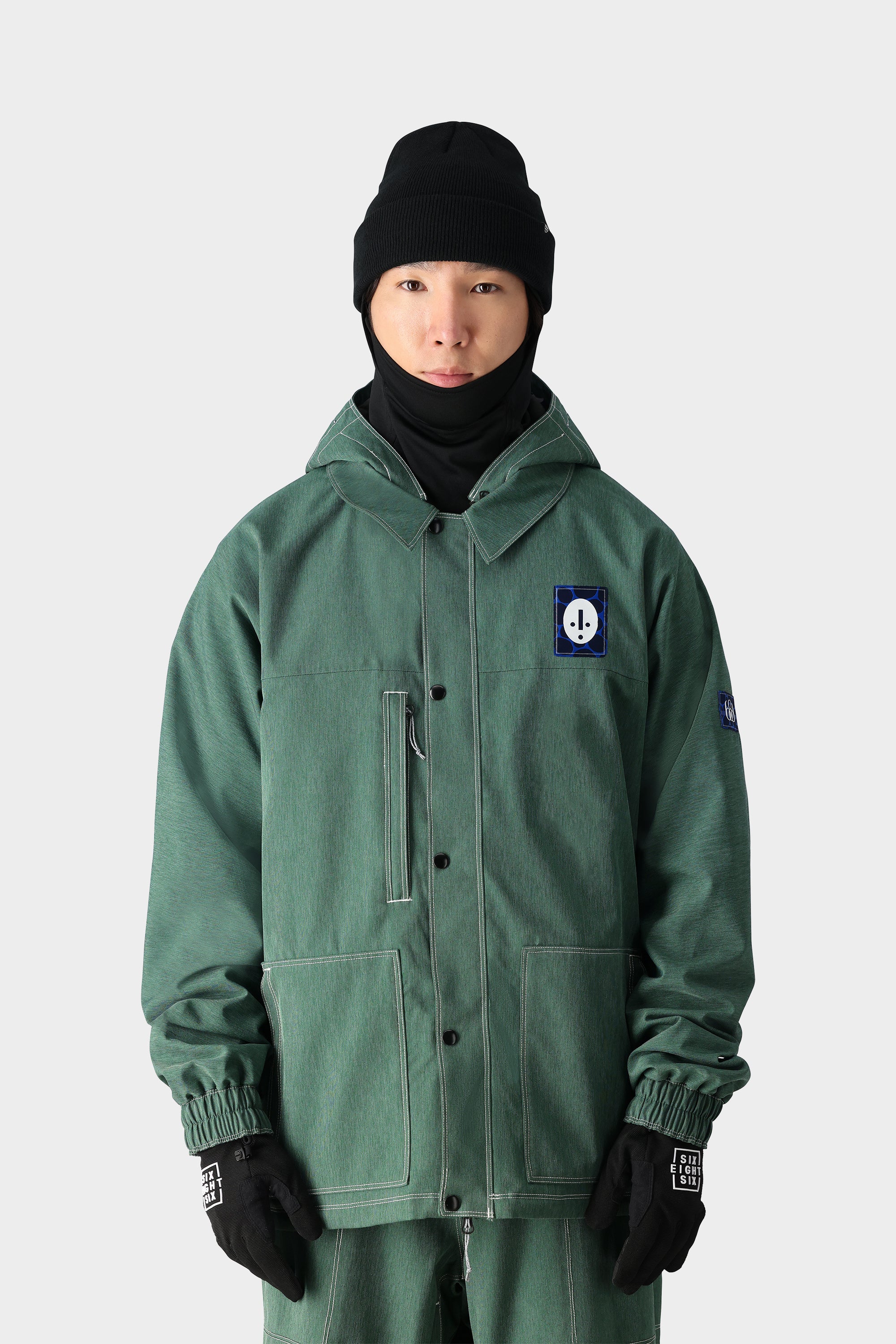 A person wearing the 686 Dojo Jacket by 686 in green, featuring a blue and white patch, along with black gloves and a black beanie, stands facing forward against a plain light gray background.