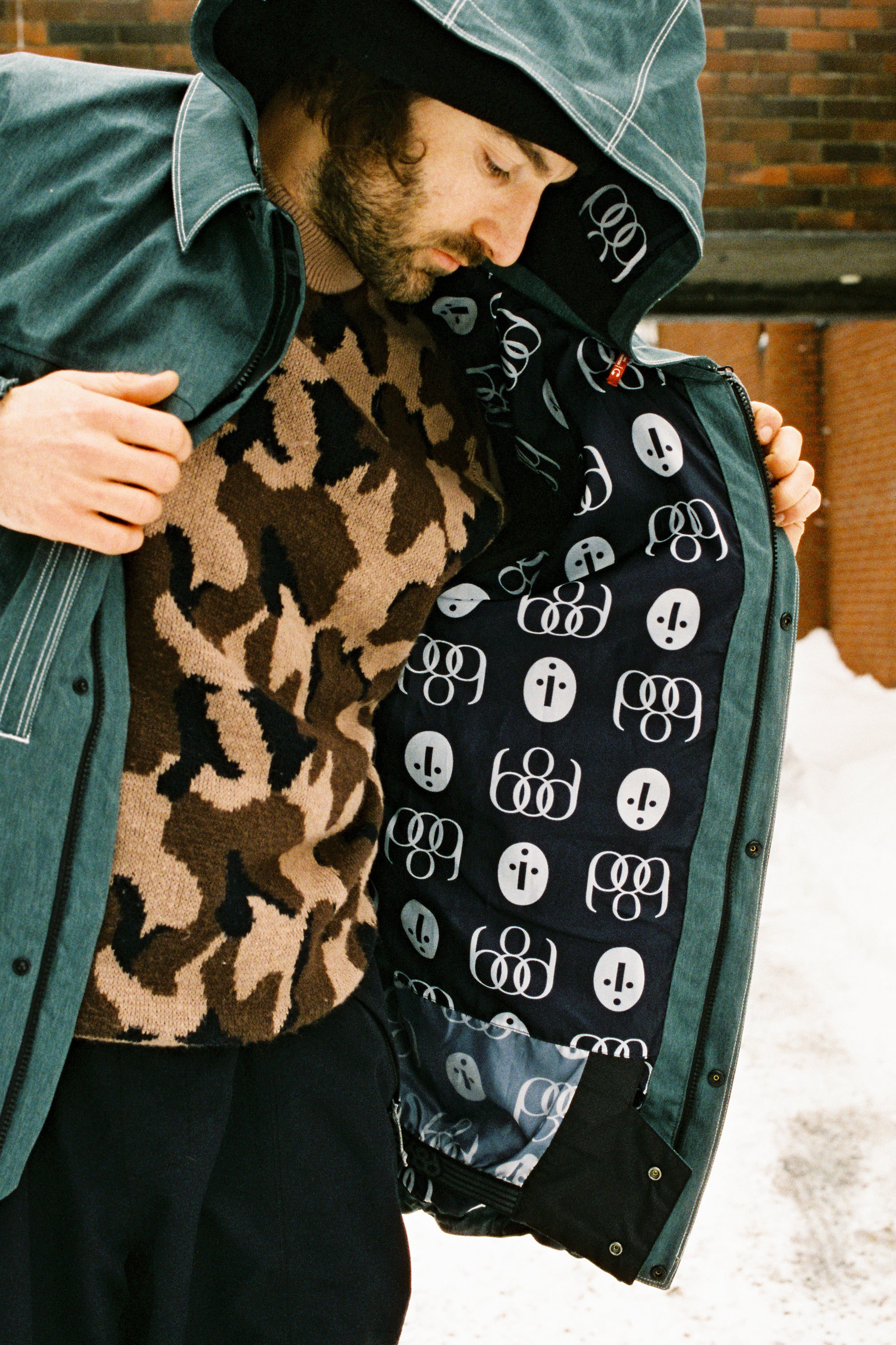 A person wearing the 686 Dojo Jacket by 686 opens it to show its black lining with white abstract patterns. They pair it with a brown and black patterned sweater and stand outdoors by a brick wall on snowy ground.