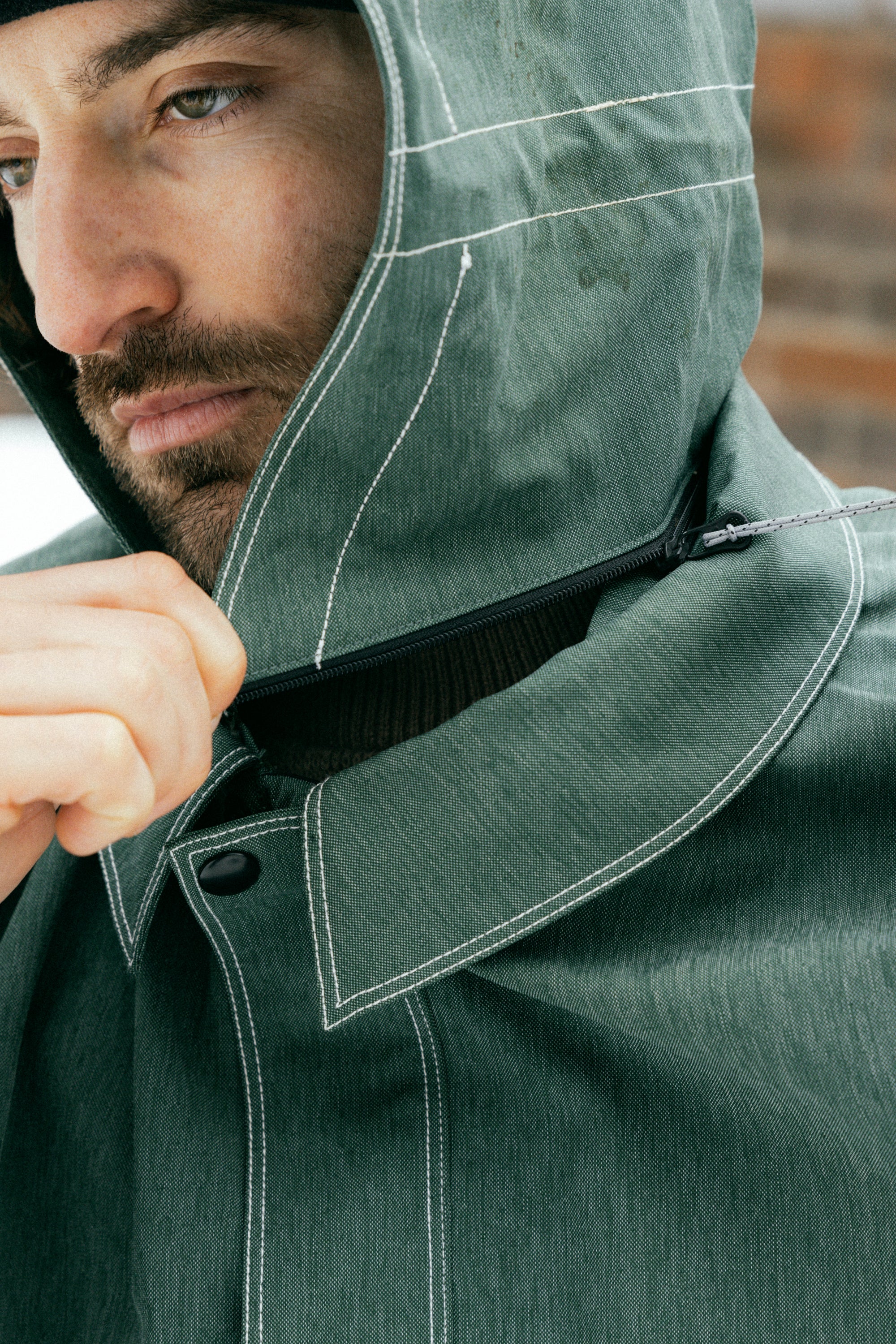 A close-up of a bearded person in the 686 Dojo Jacket by 686, featuring green fabric, white stitching, and technical details inspired by Forest Bailey. The individual zips up the front while looking down; background is blurred.