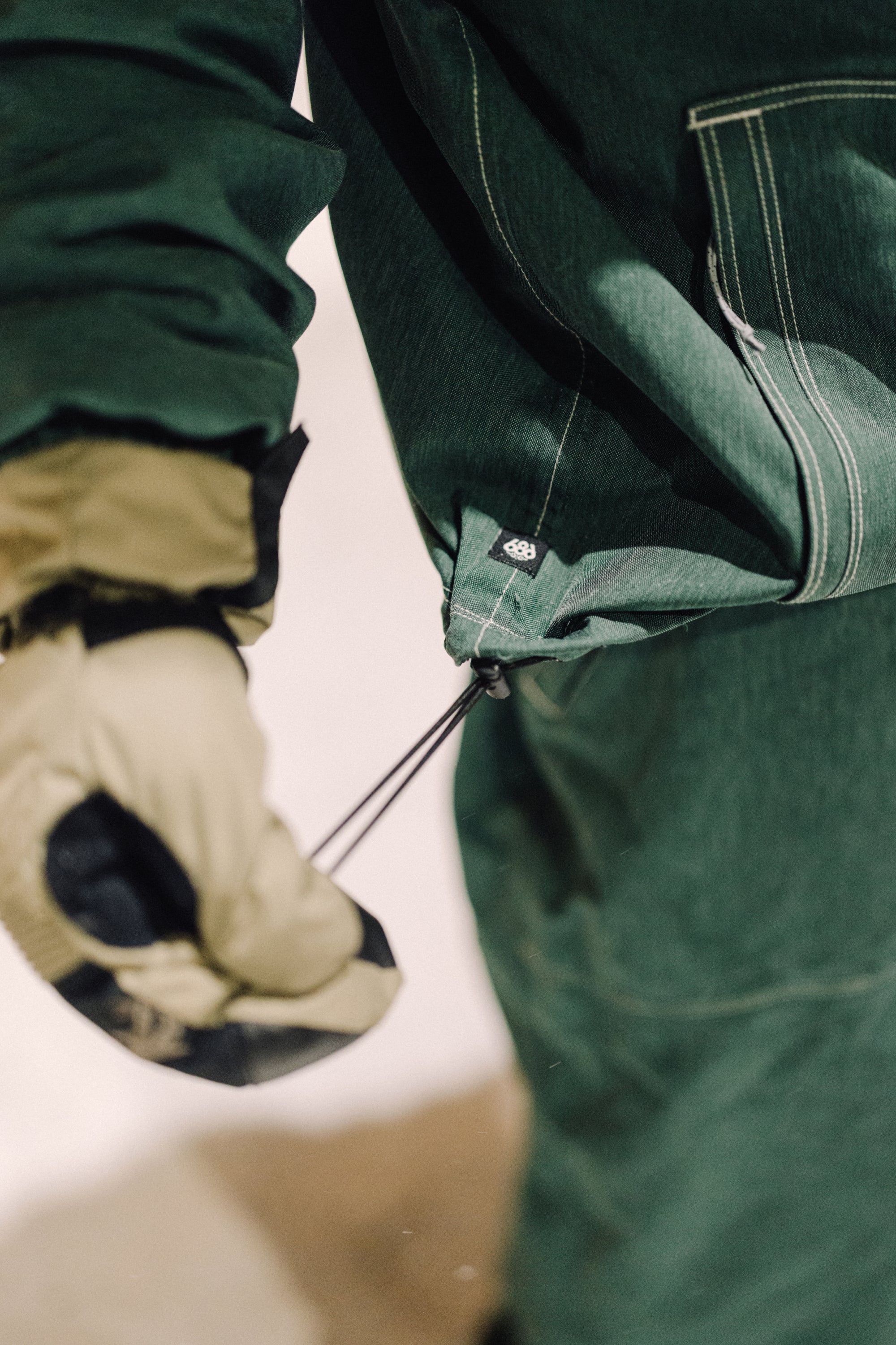 A person wearing a green 686 Dojo Jacket and tan glove tightens the elastic drawcord at the cuff for a secure fit, showcasing technical features inspired by Forest Bailey.