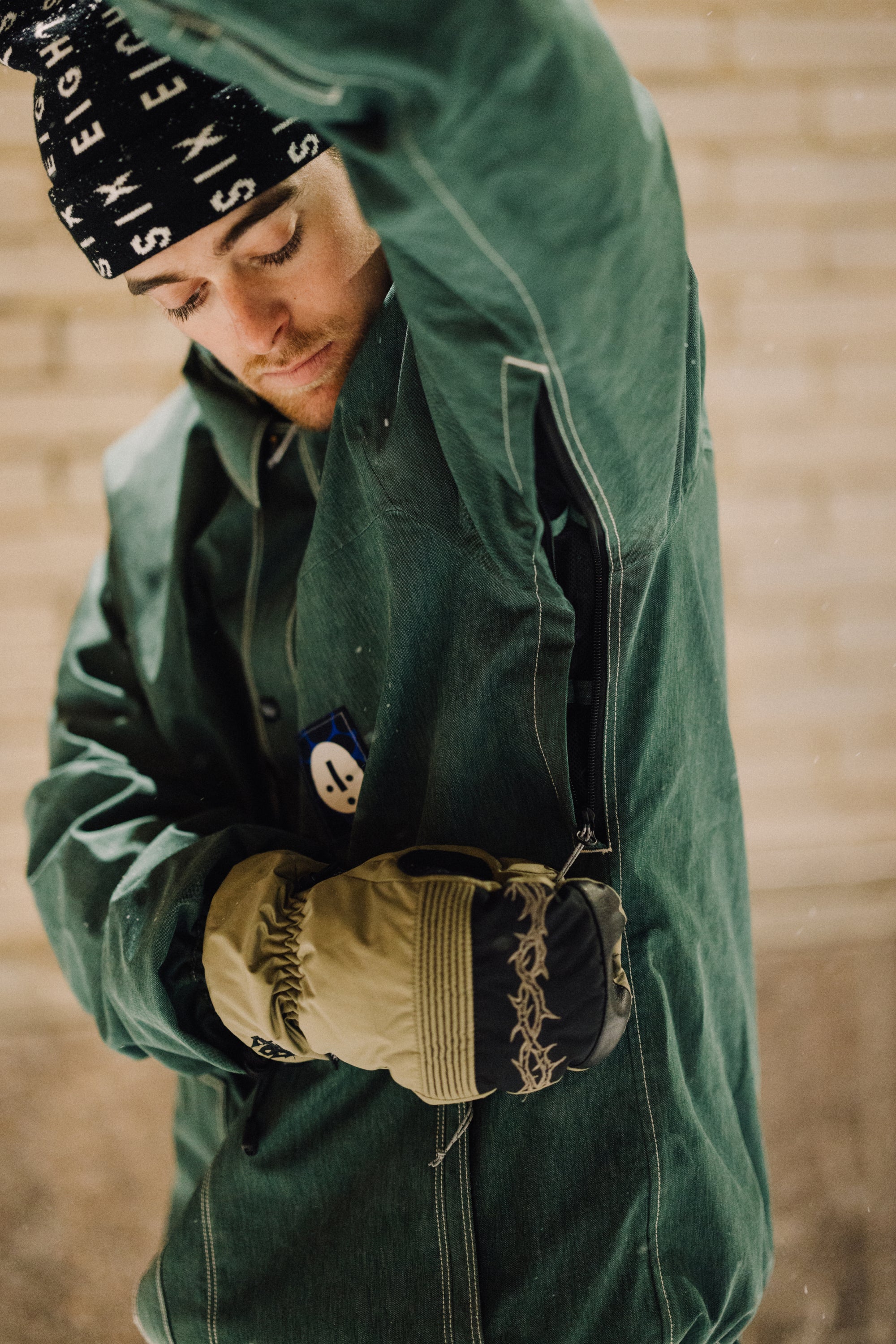 A person in the green 686 Dojo Jacket with technical features, patterned gloves, and a black beanie unzips under the arm, head lowered and eyes closed. A beige brick wall forms the background.