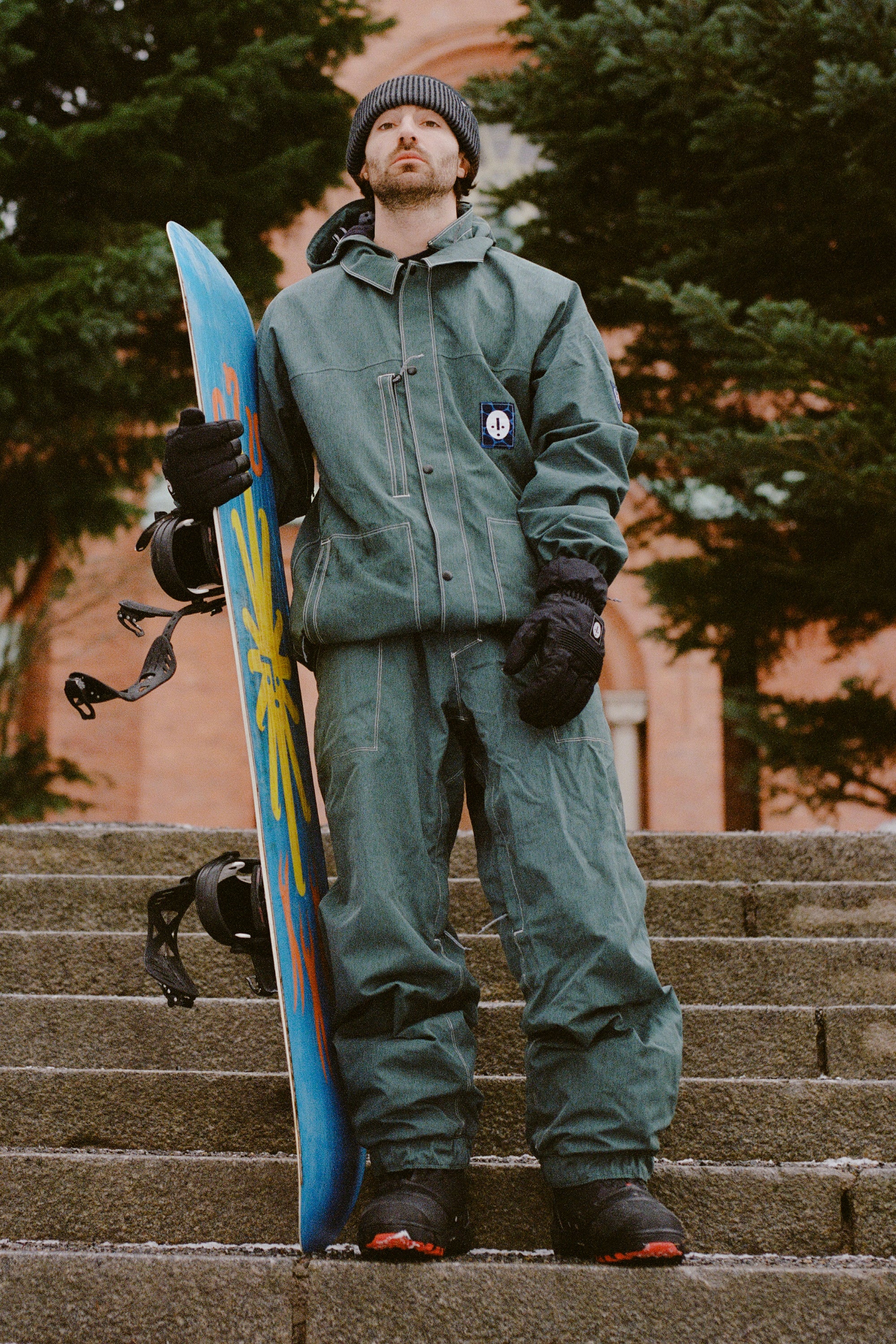 Wearing the 686 Dojo Jacket and a beanie, a person stands on outdoor steps holding a bright snowboard upright, with trees and a brick building behind them—reflecting Forest Bailey’s bold style.