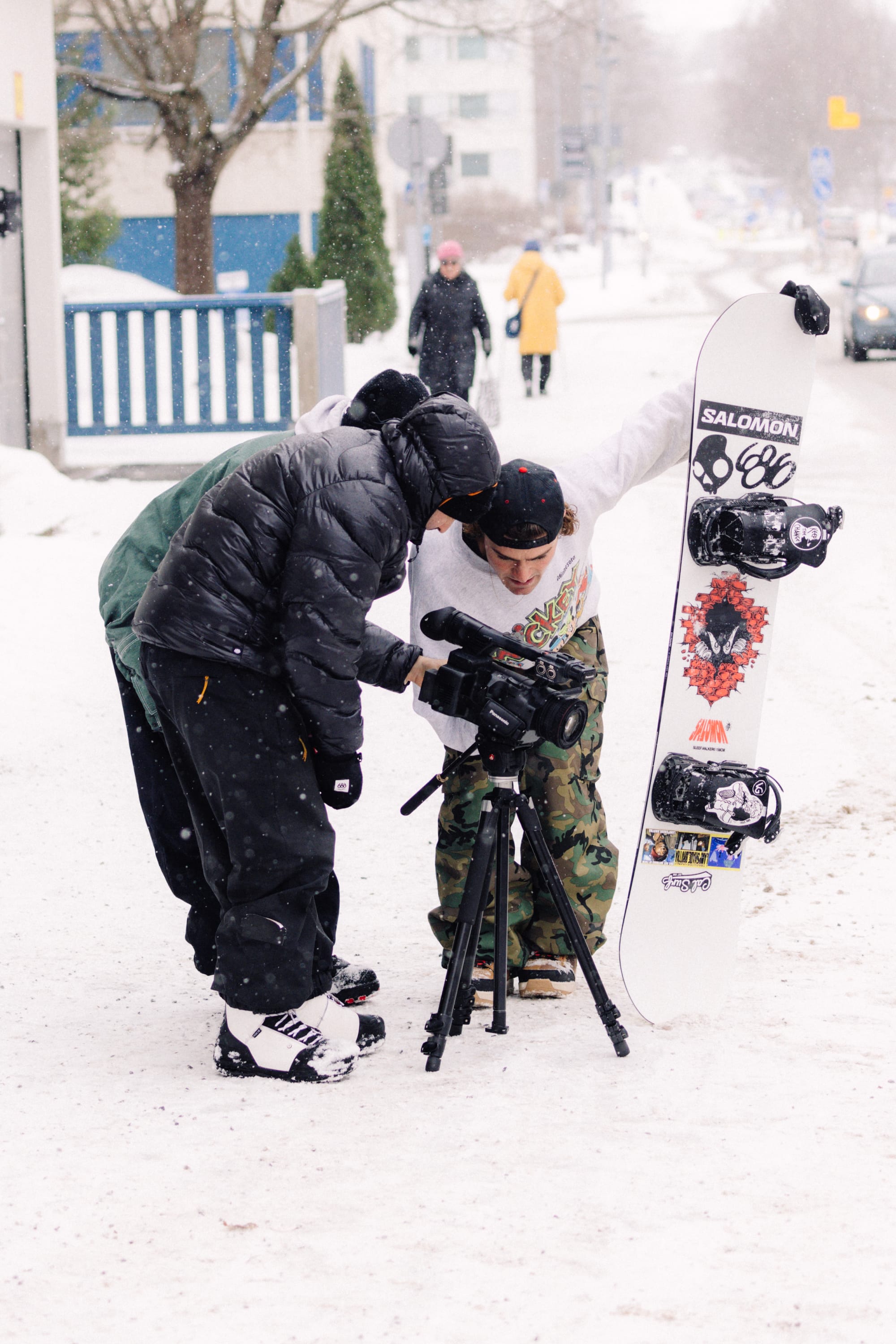 Three people stand in the snow—a pair examining a video camera mounted on a tripod, while another holds a snowboard upright—wearing the 686 Men’s Essox™ Cargo Pant by 686. Snow-dusted buildings and a passing pedestrian appear in the background.