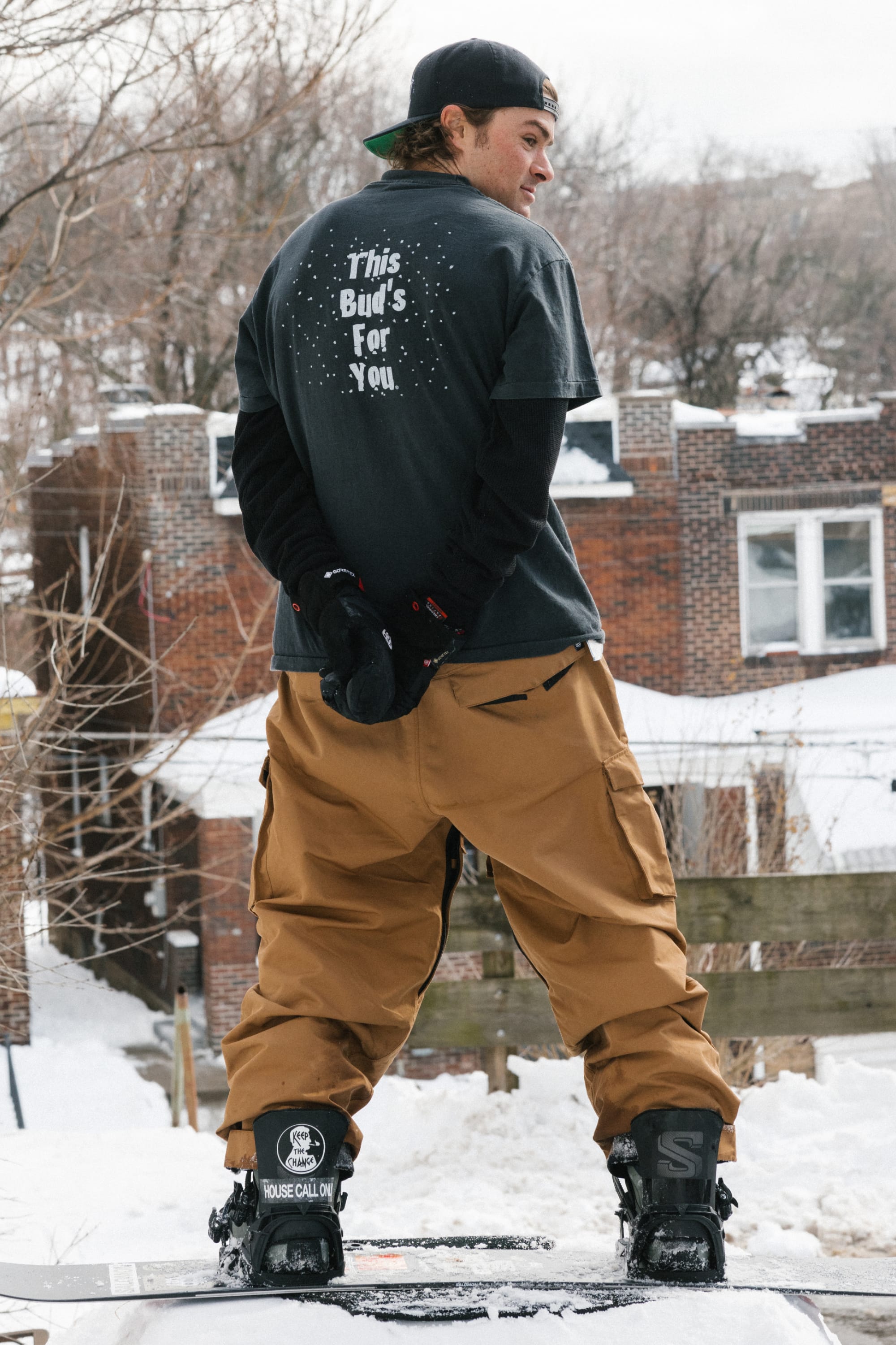 A person in a black t-shirt and 686 Men’s Essox™ Cargo Pant by 686 stands on a snowboard in the snow, facing away. Their shirt reads “This Buds For You.” Brick houses and bare trees appear in the background.
