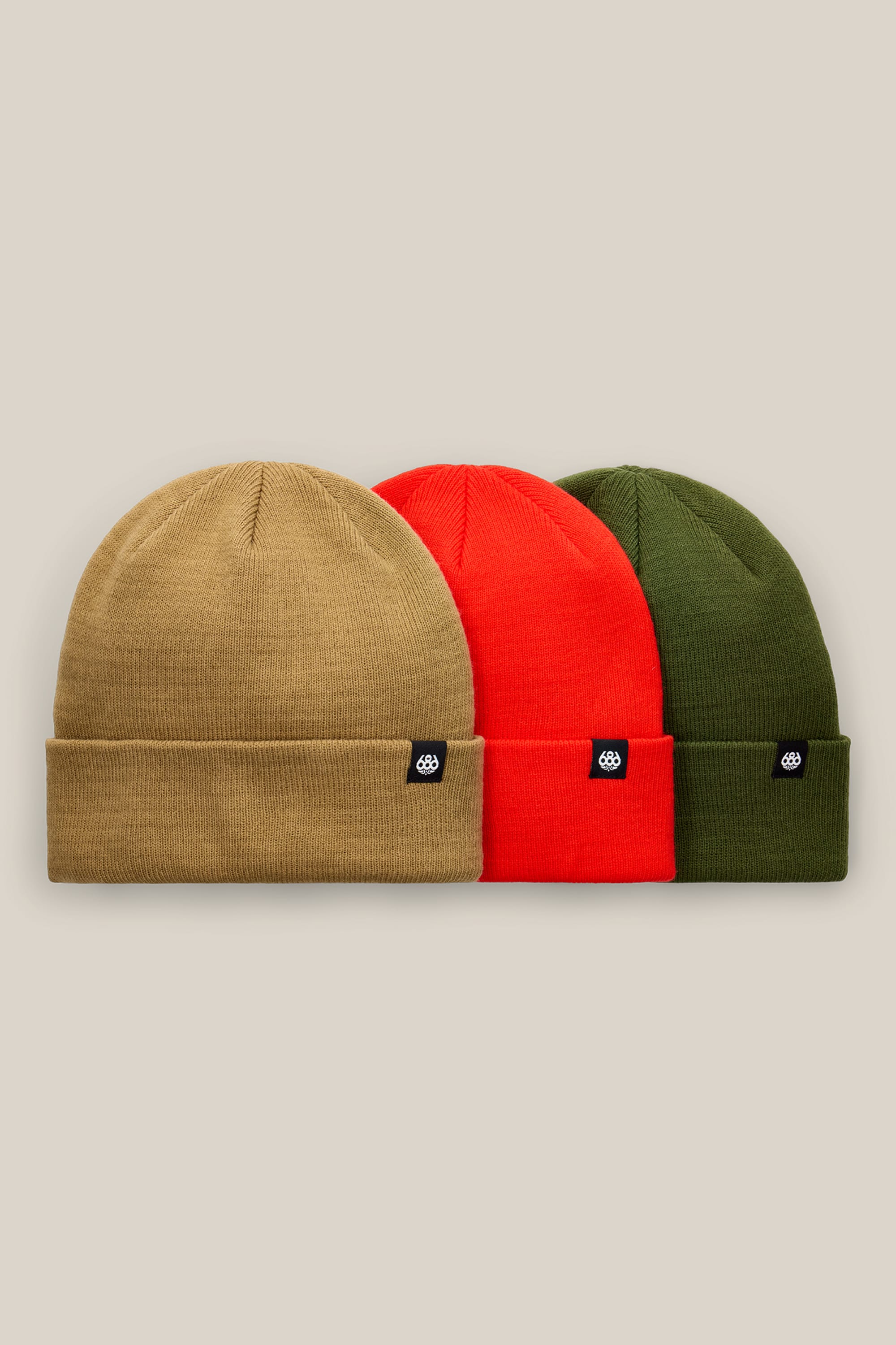 The 686 Standard Roll-Up Beanie 3-Pack by 686 features tan, red, and olive green knit beanies, each adorned with a black rectangular patch displaying a white logo, lined up side by side against a beige background.