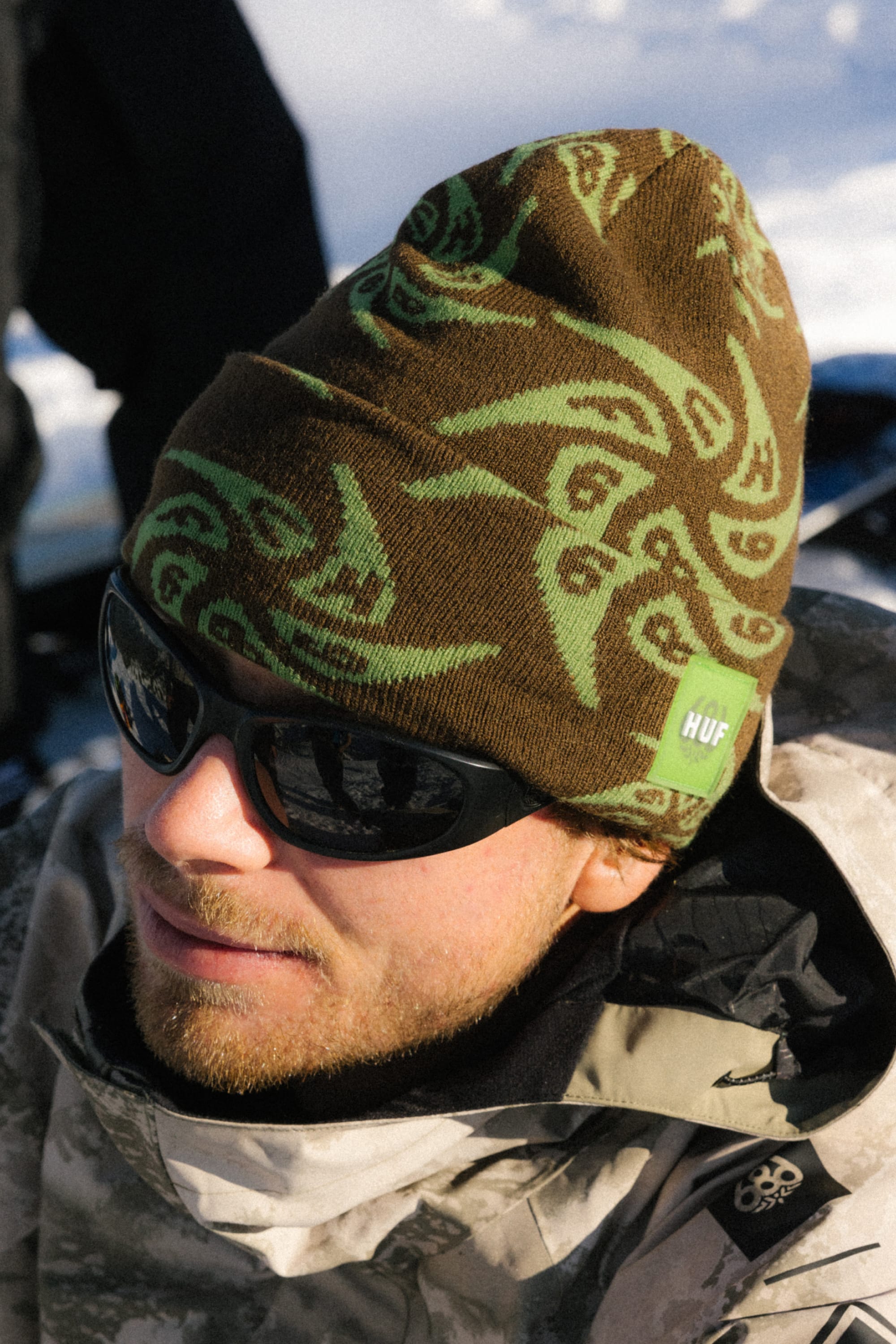 A person wearing black sunglasses, a camo-pattern jacket, and a brown and green 686 HUF Beanie from 686 stands outdoors in the snow.