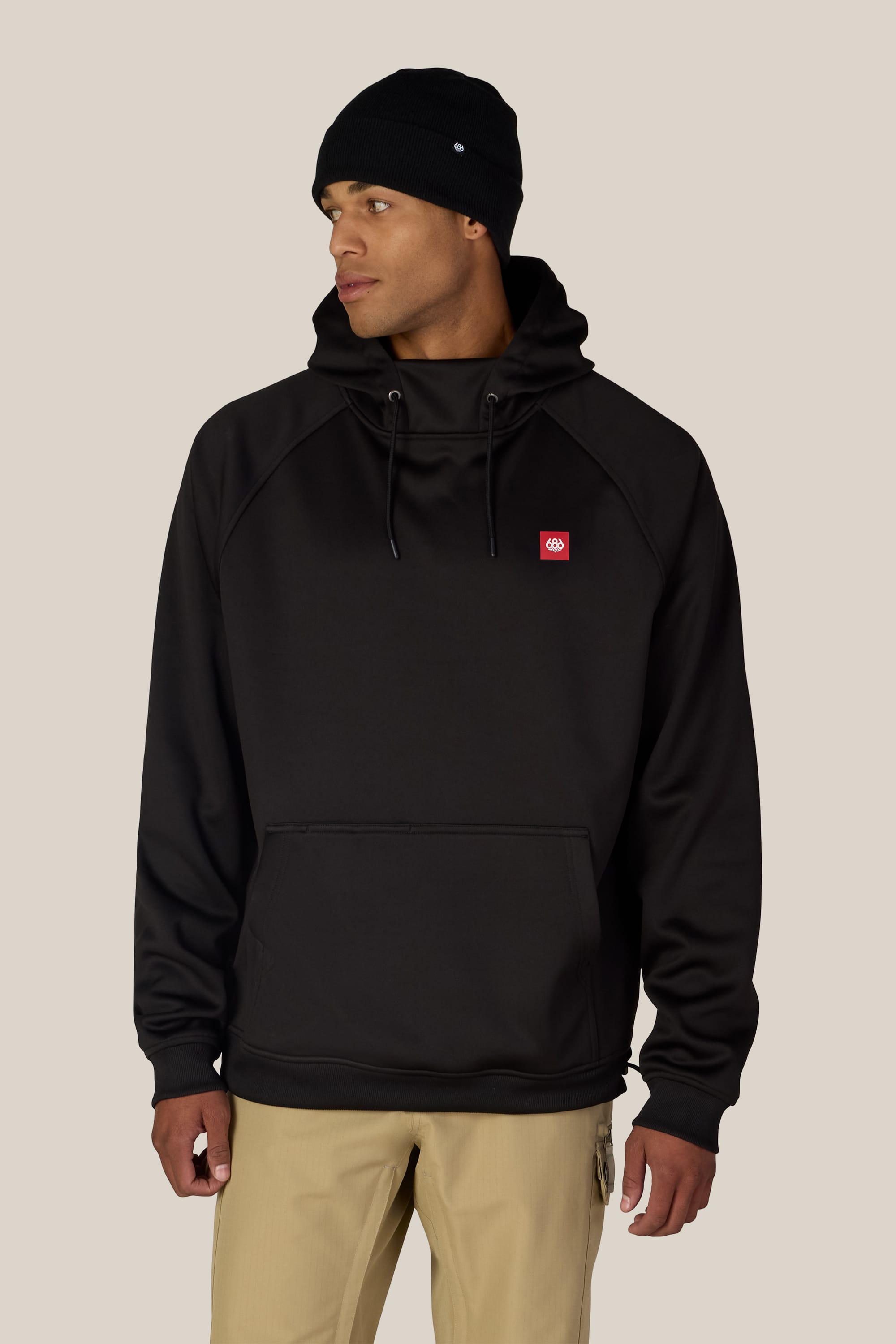 A person wearing a black beanie and the 686 Men’s Bonded Fleece Pullover Hoody by 686, paired with beige pants, stands against a light beige background, looking to the side.