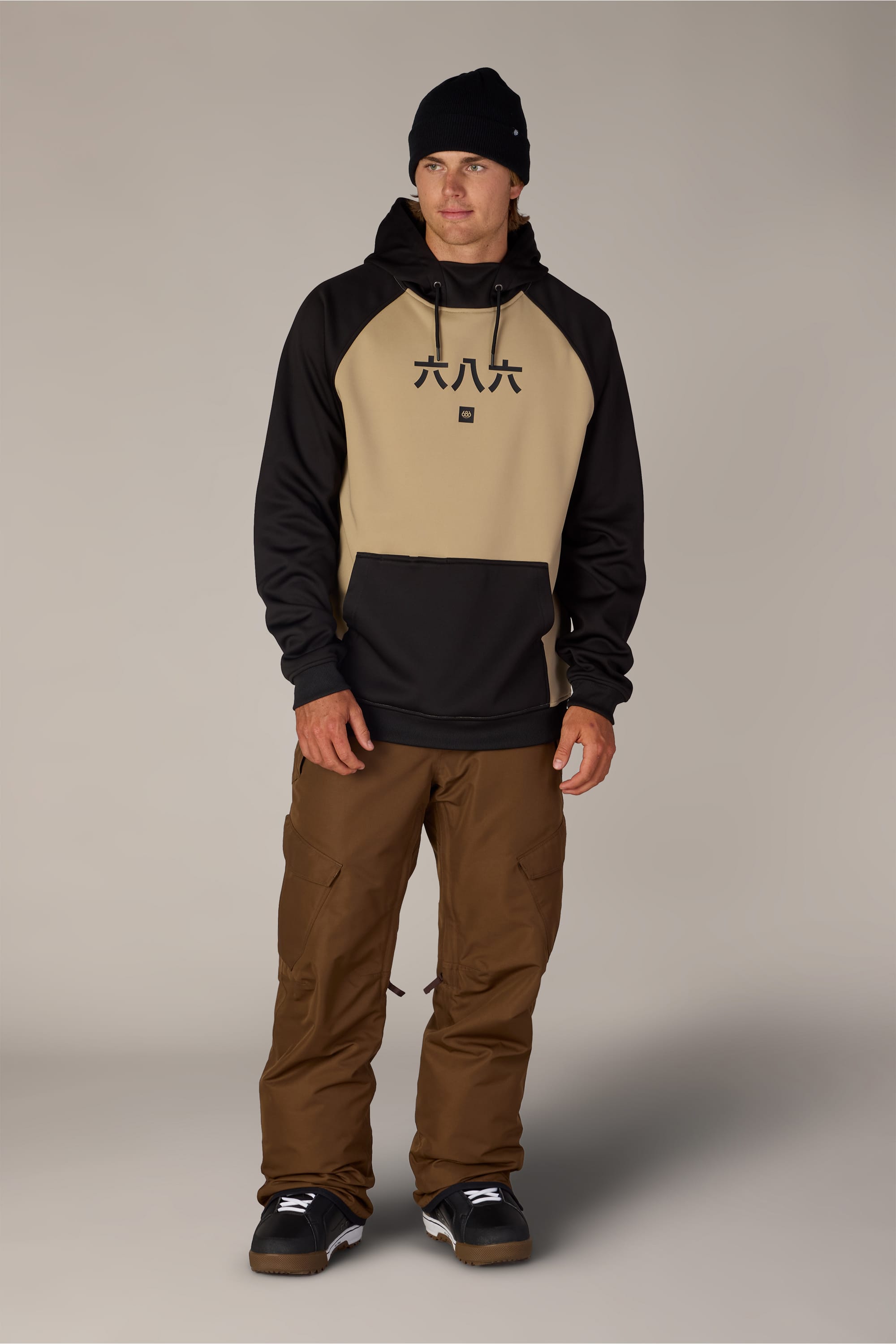 A man wearing a black beanie, the 686 Men’s Bonded Fleece Pullover Hoody in tan and black, brown pants, and black sneakers stands against a plain background, facing forward with a neutral expression.