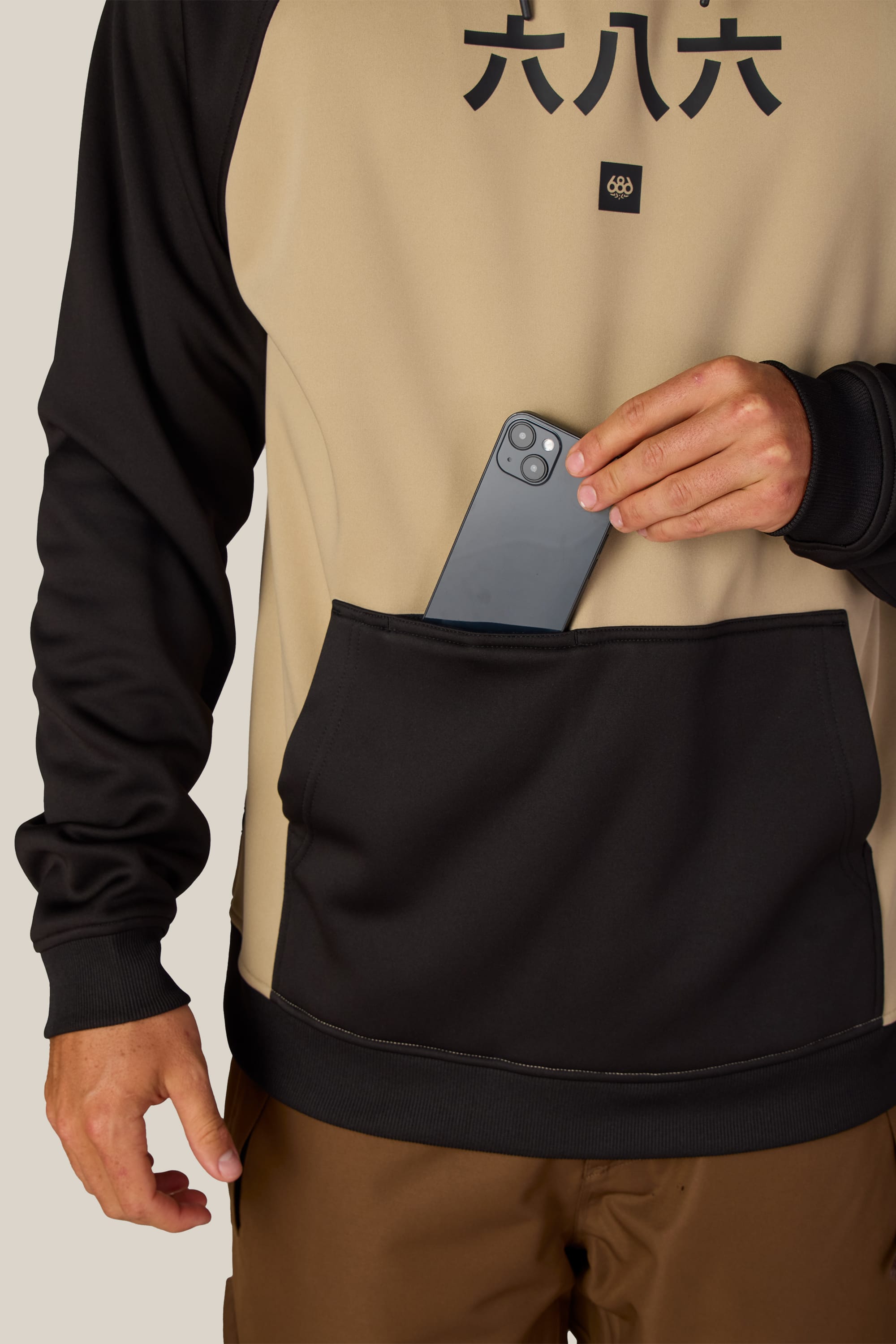 A person wearing the 686 Men’s Bonded Fleece Pullover Hoody in beige and black places a smartphone into its front pocket. Only the lower half of their face and torso are visible.