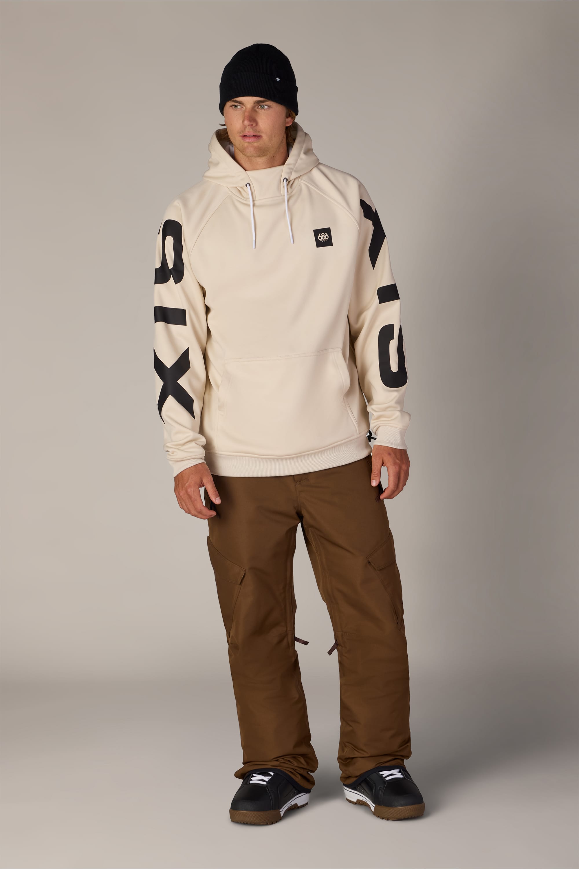 A man wearing the 686 Men’s Bonded Fleece Pullover Hoody in cream, featuring “SIX” in black on both sleeves, along with brown pants, black sneakers, and a black beanie stands against a plain light background.
