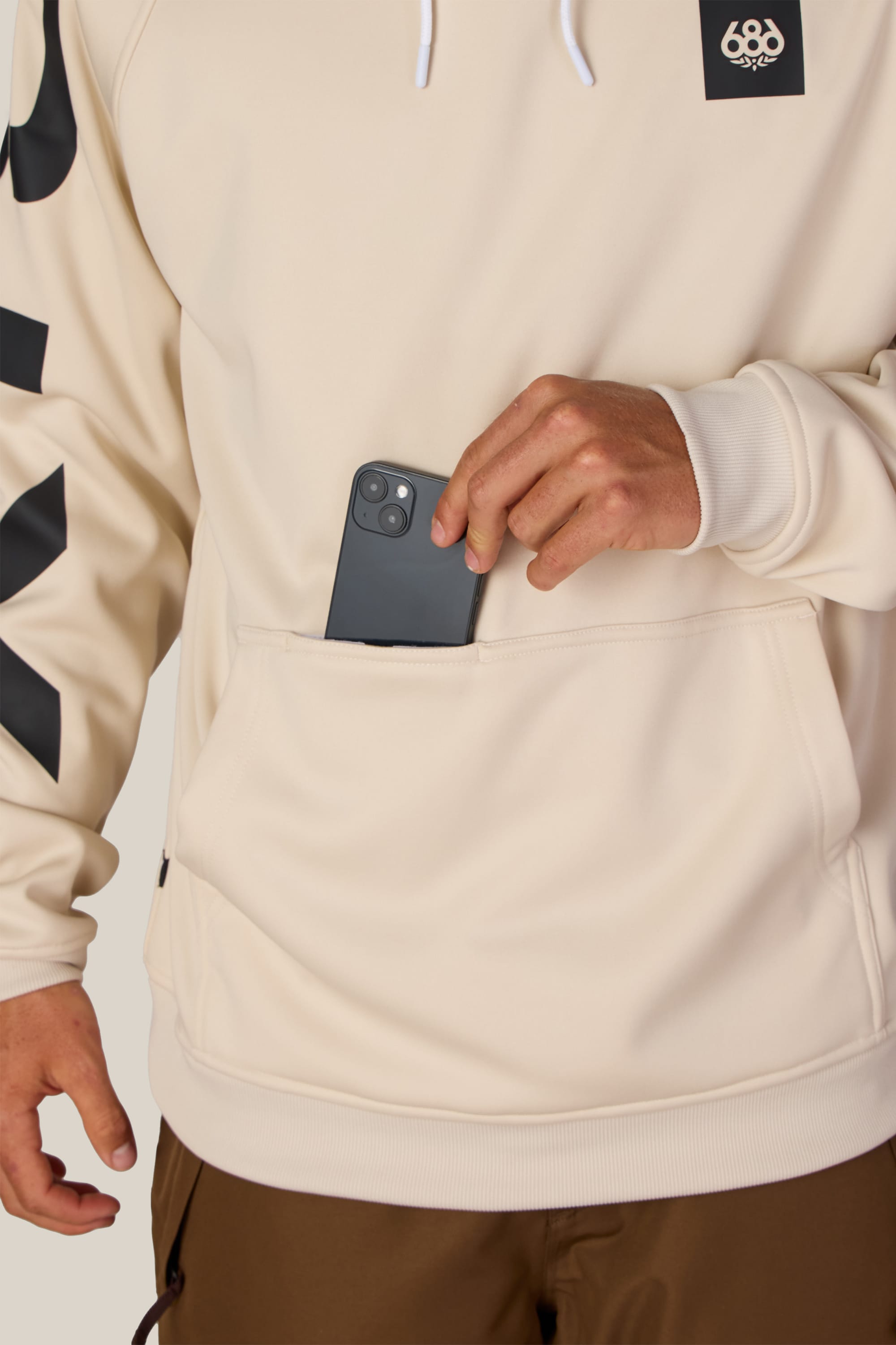 A person wearing the 686 Men’s Bonded Fleece Pullover Hoody in cream with black graphics is placing a black smartphone into the front pocket. Only their torso and one hand are shown.