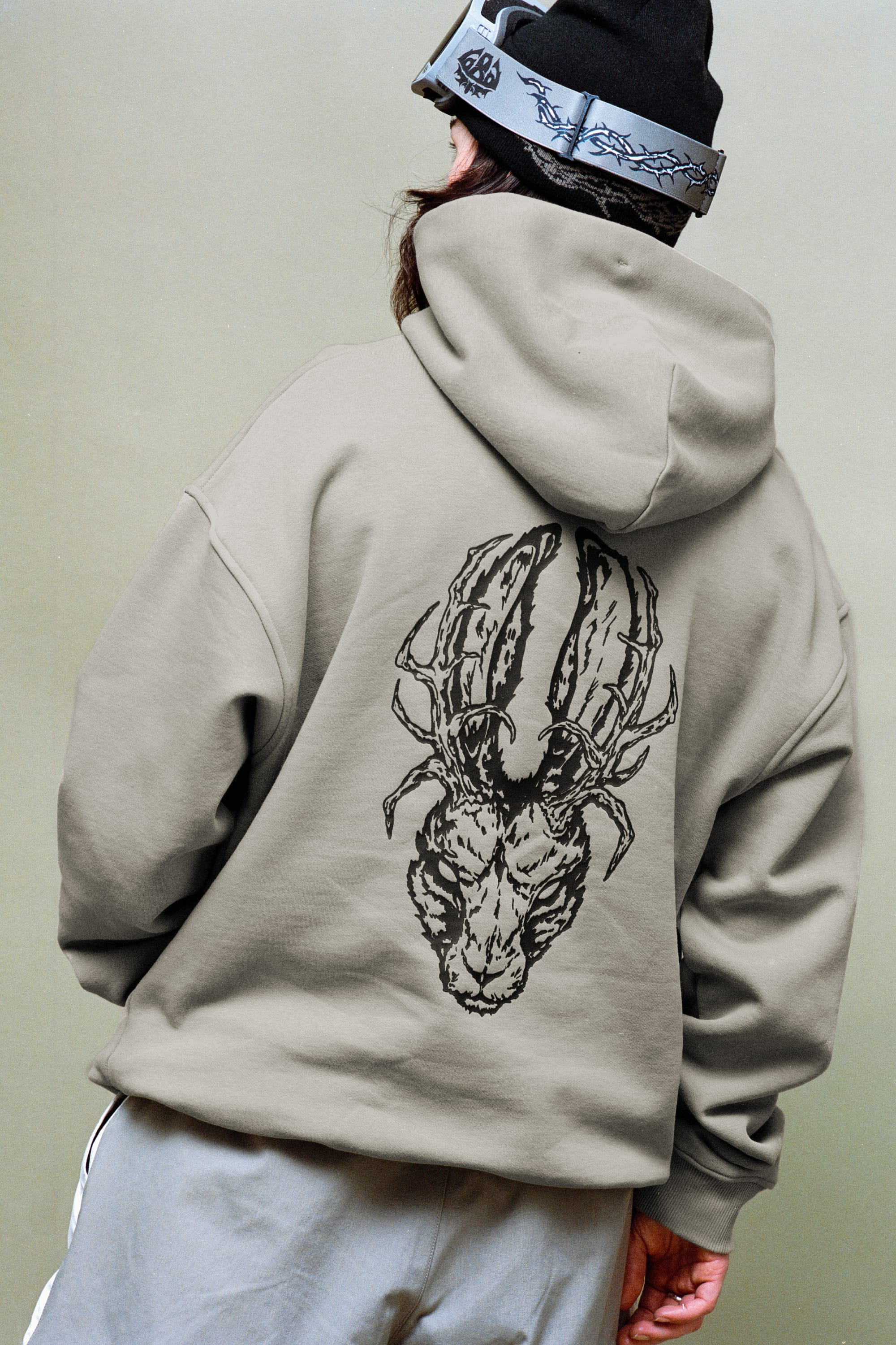A person faces away, wearing the 686 Mens Welcome Premium Heavyweight Pullover Hoody in taupe with a bold black ram’s head graphic on the back, paired with light gray pants and a black beanie with goggles for a boxy, street-ready look.