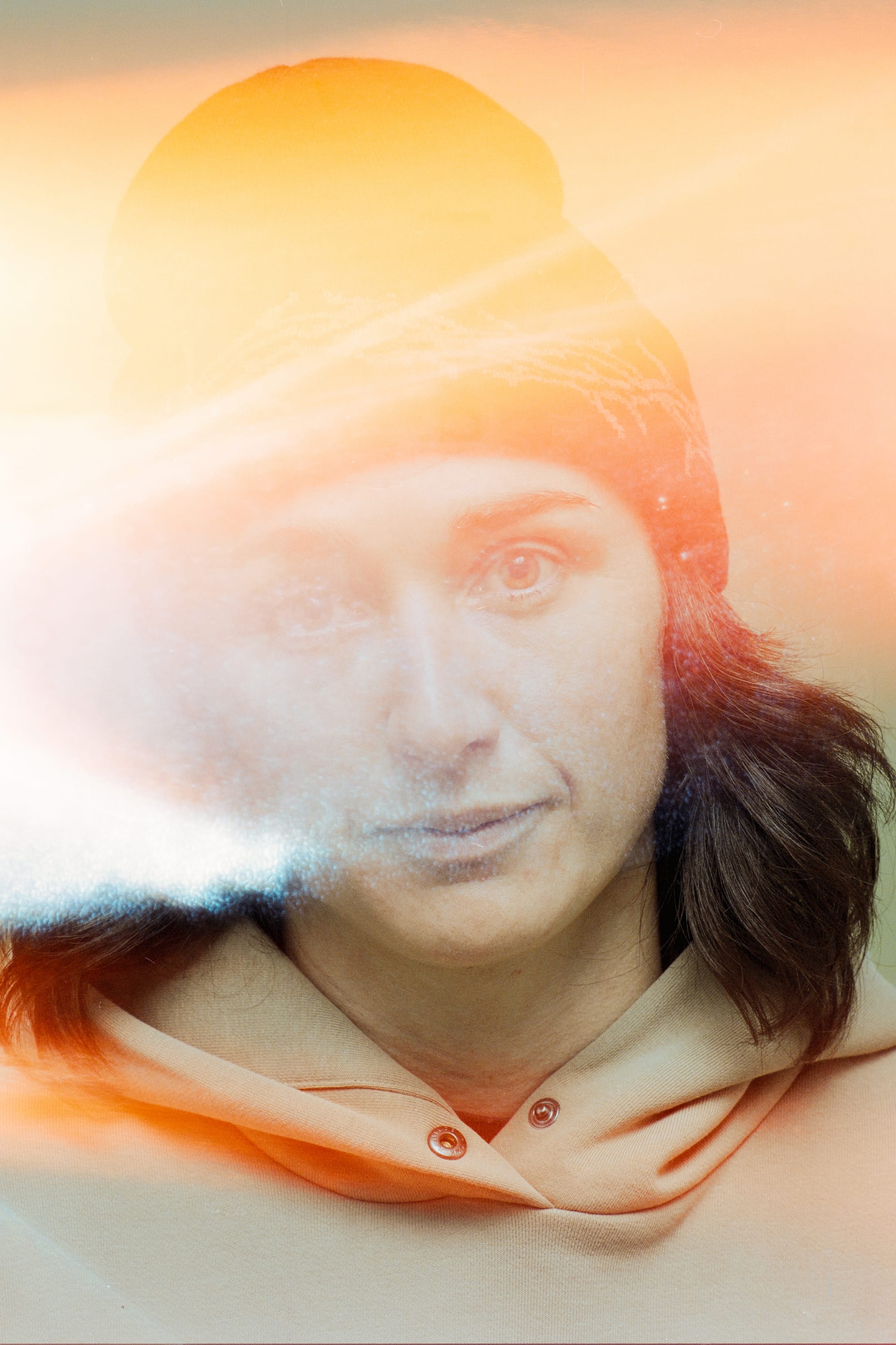 A person with shoulder-length brown hair, wearing a 686 Welcome Beanie by 686 and a beige hoodie, looks directly at the camera as bright orange and white light leaks partially obscure their face.