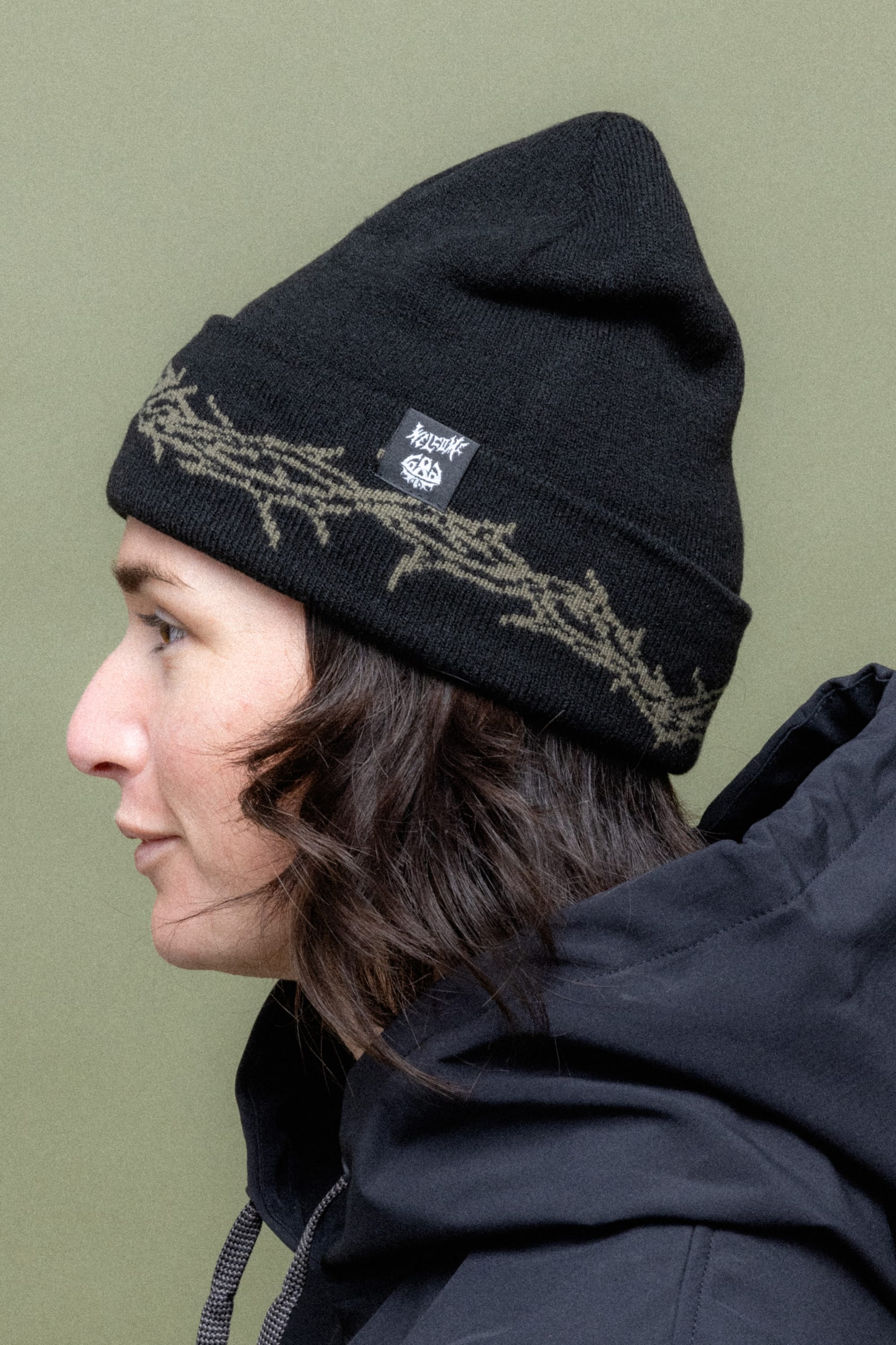 A person with wavy, dark hair in profile wears a 686 Welcome Beanie by 686, featuring a woven branch pattern and a small patch, along with a dark jacket, set against an olive green background.