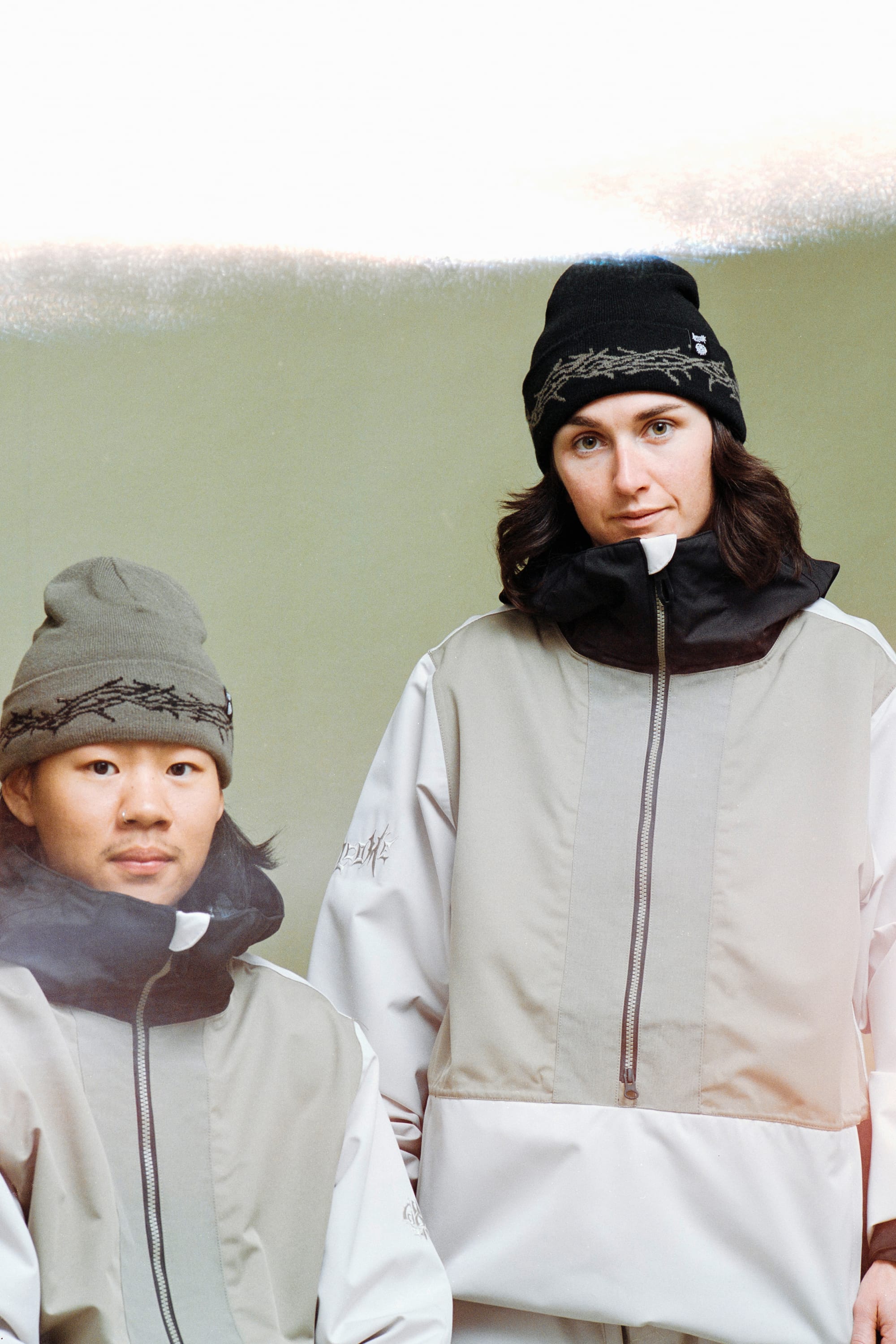 Two people in matching light grey jackets with black zippers and 686 Welcome Beanie hats by 686 pose together against a plain background. One is seated, the other stands beside them, both facing the camera.