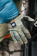 A person wearing a green camo 686 Mens Outlaw Pipe Glove by 686 with enhanced grip and a teal jacket sleeve stands beside a car on cracked pavement.