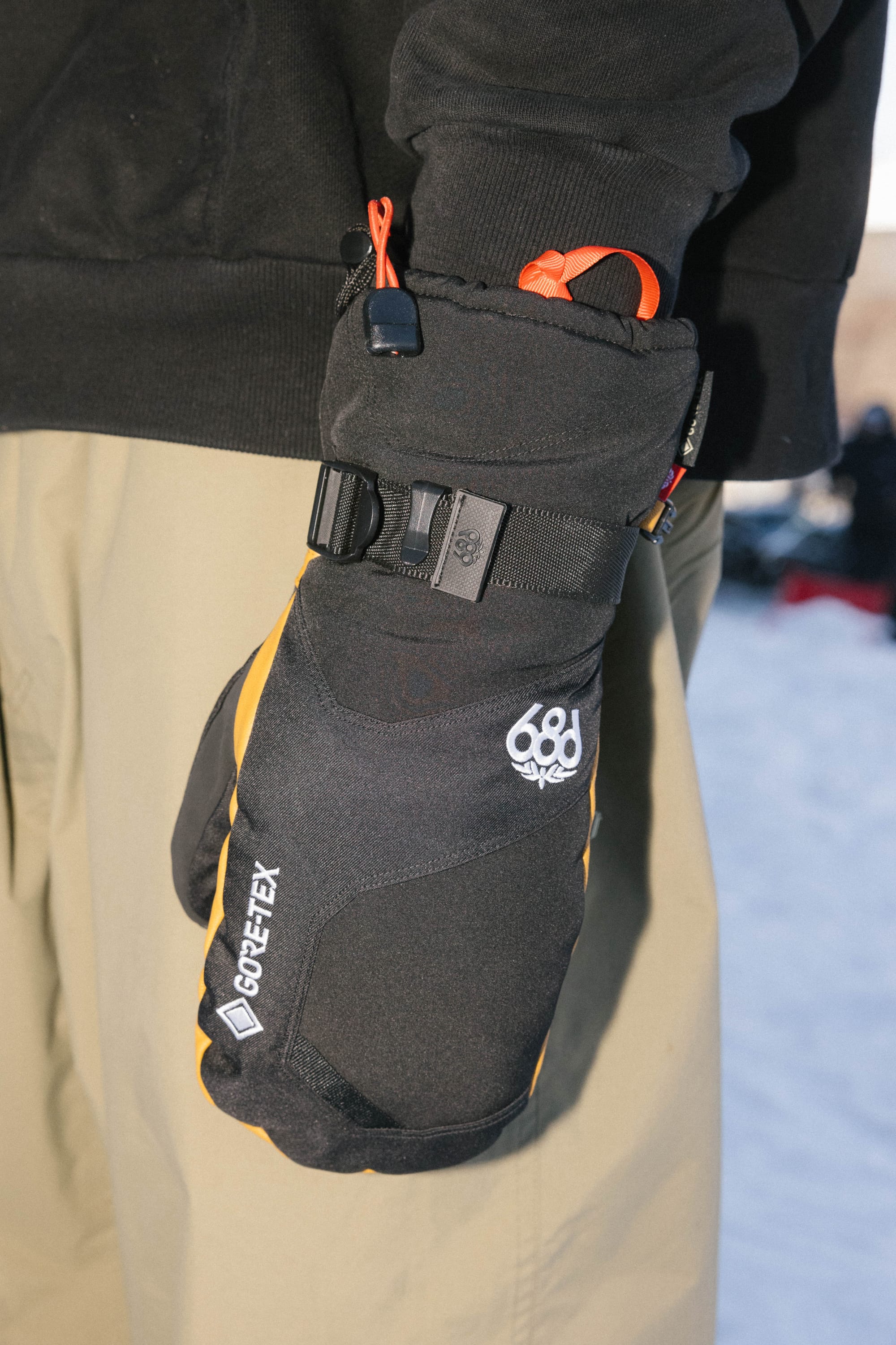 A person wearing tan pants, a black jacket, and the 686 Men’s GORE-TEX® Leather Linear™ Mitts with orange accents and the 686 logo stands outdoors in a snowy area.