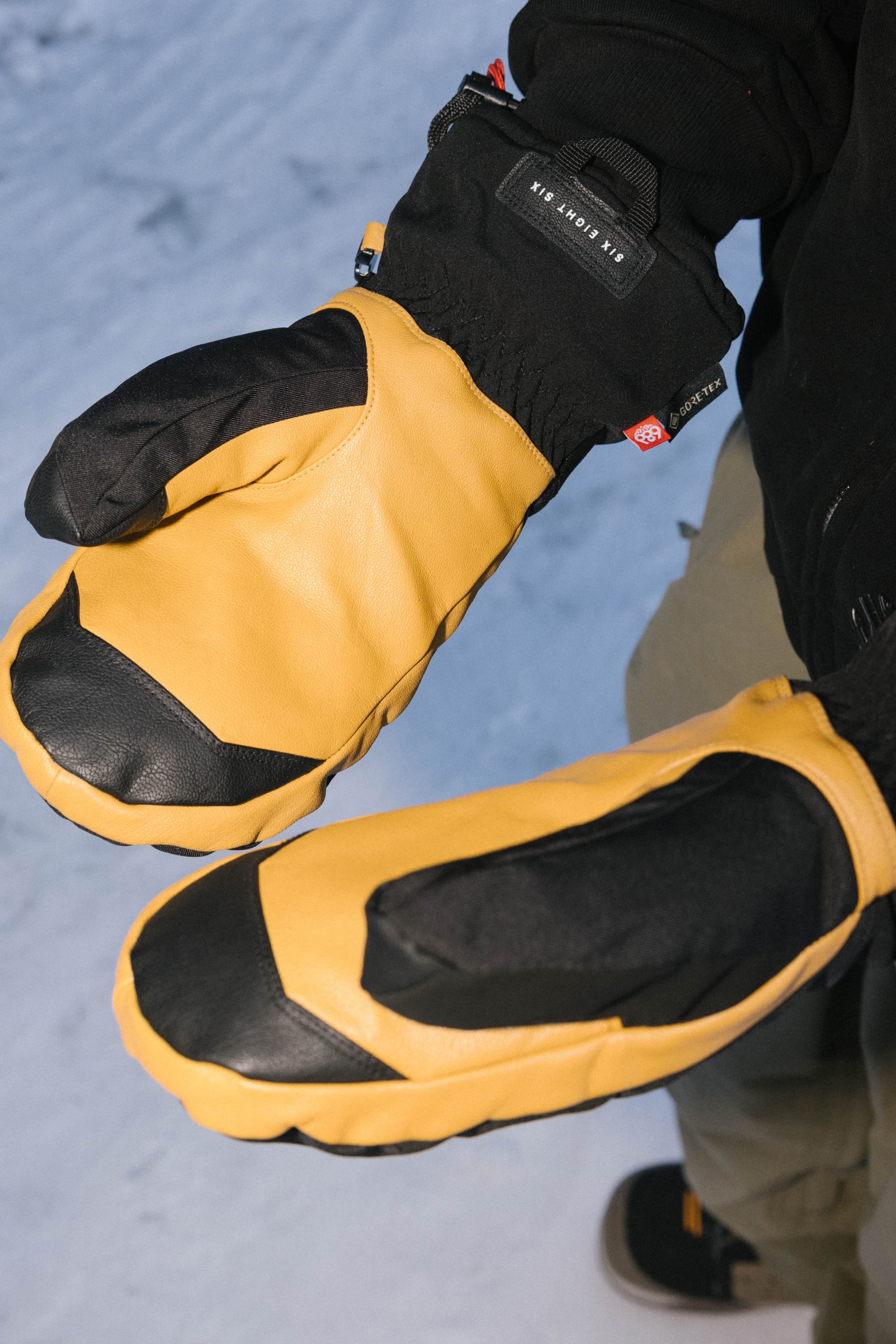 A person wearing the 686 Men’s GORE-TEX® Leather Linear™ Mitt by 686 stands on snow, hands extended forward to display the mittens’ design and textured material.
