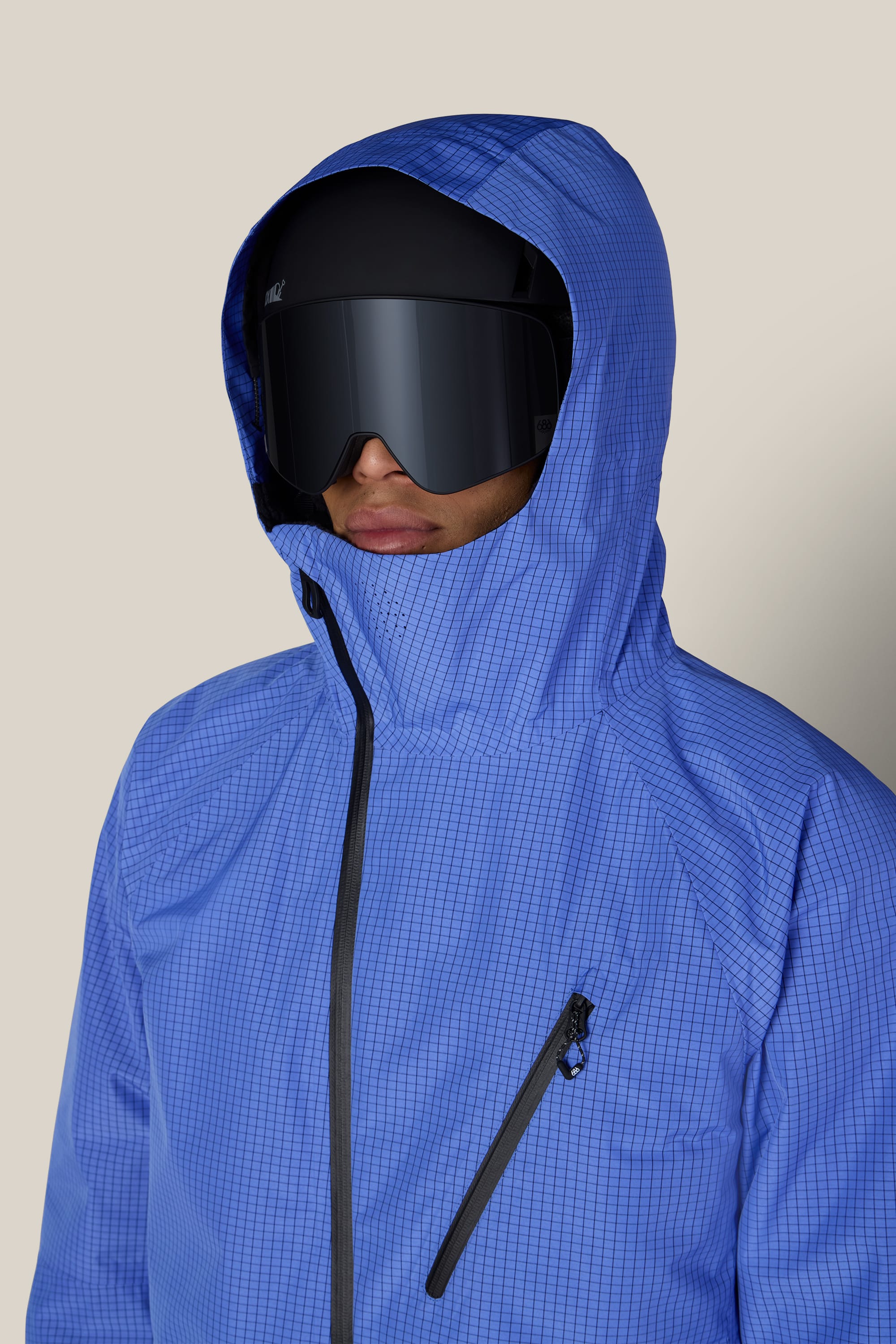A person wearing the 686 Men’s Hydra™ Thermagraph® Jacket by 686—a blue, grid-patterned hooded jacket featuring a slanted chest zipper—along with large black visor sunglasses, is facing slightly to the side against a plain beige background.