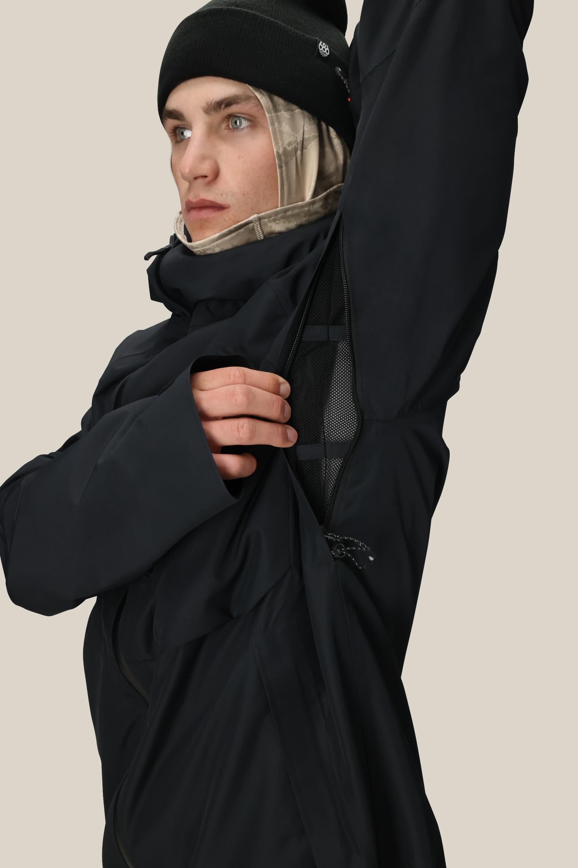Wearing the 686 Men’s Hydra™ Thermagraph® Jacket, a person in a black jacket and beanie lifts their arm to reveal a mesh-lined zipper vent under the armpit, showcasing the jacket’s advanced ventilation feature from 686.