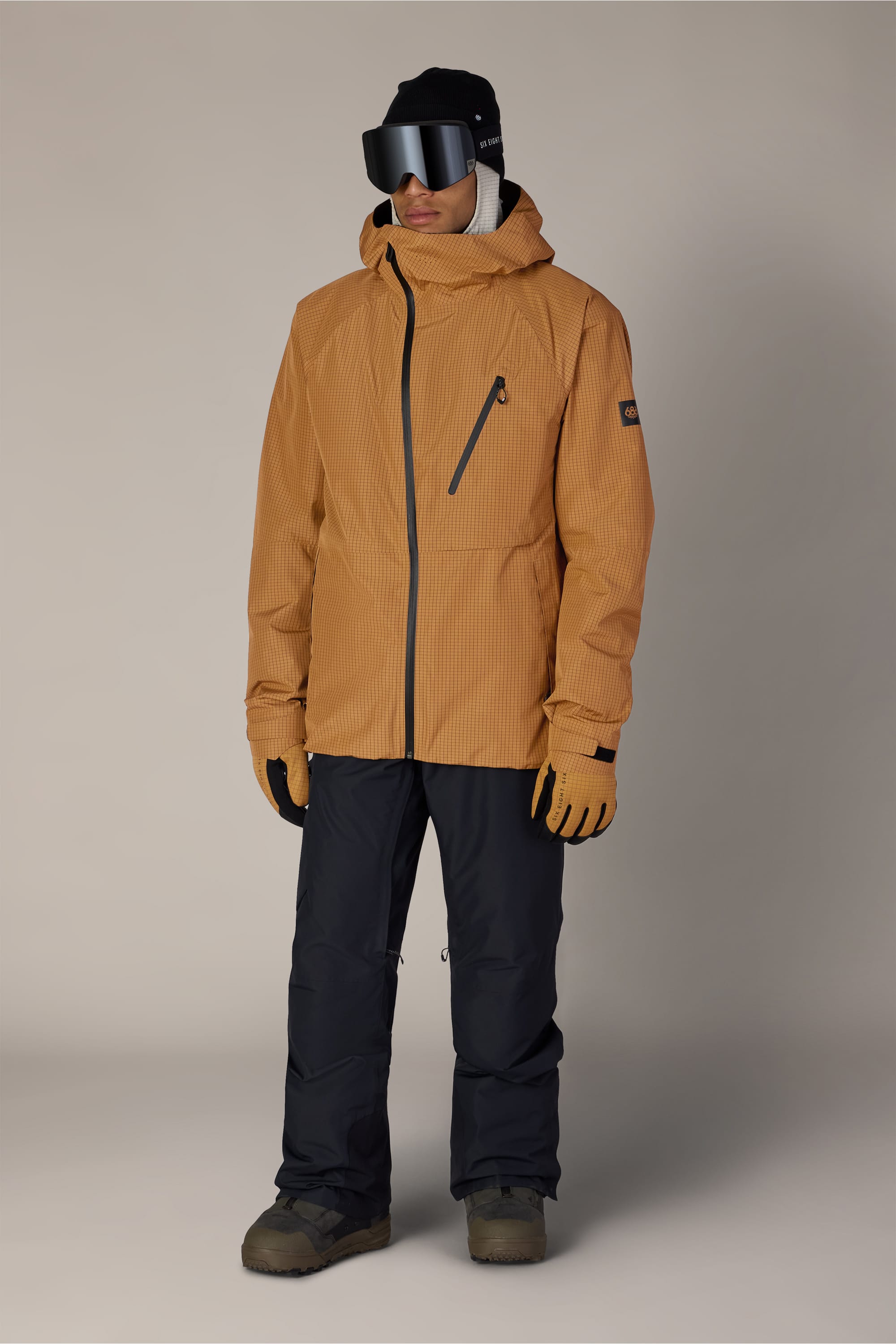 A person wearing the 686 Men’s Hydra™ Thermagraph® Jacket in tan, along with black snow pants, gloves, black ski goggles, and a black beanie, standing against a plain light background.