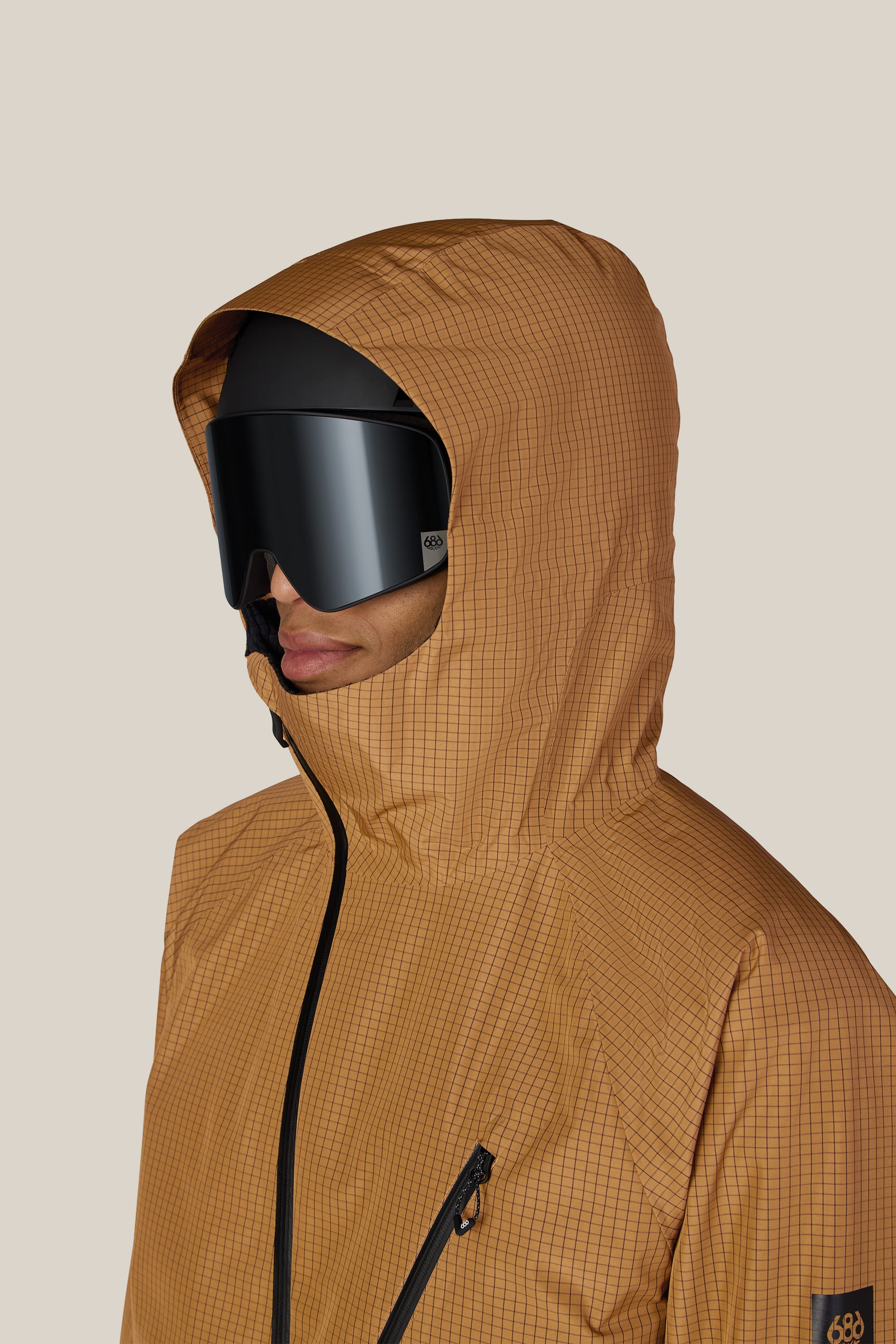 A person wearing the 686 Men’s Hydra™ Thermagraph® Jacket in a tan grid pattern and large black ski goggles, facing left against a plain light background.