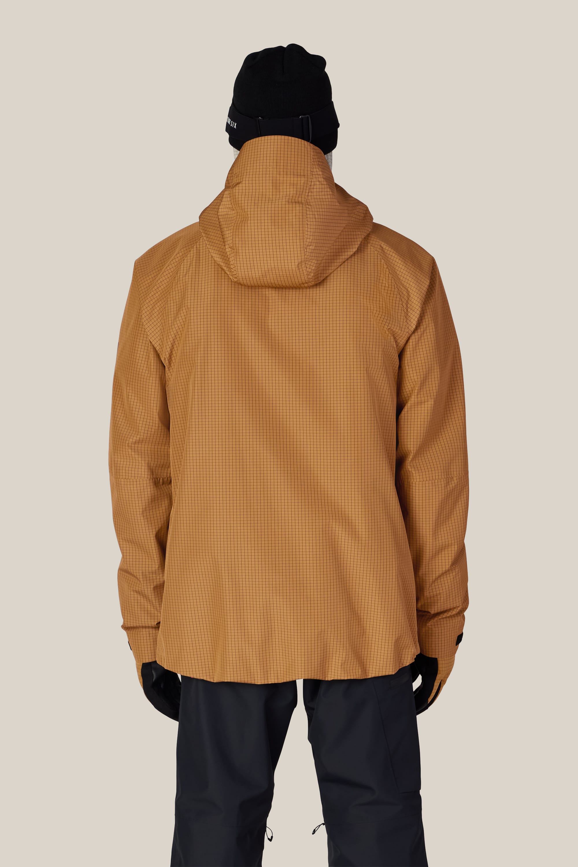 A person is shown from the back, wearing the 686 Men’s Hydra™ Thermagraph® Jacket in tan, paired with black pants, black gloves, and a black beanie, standing against a plain beige background.