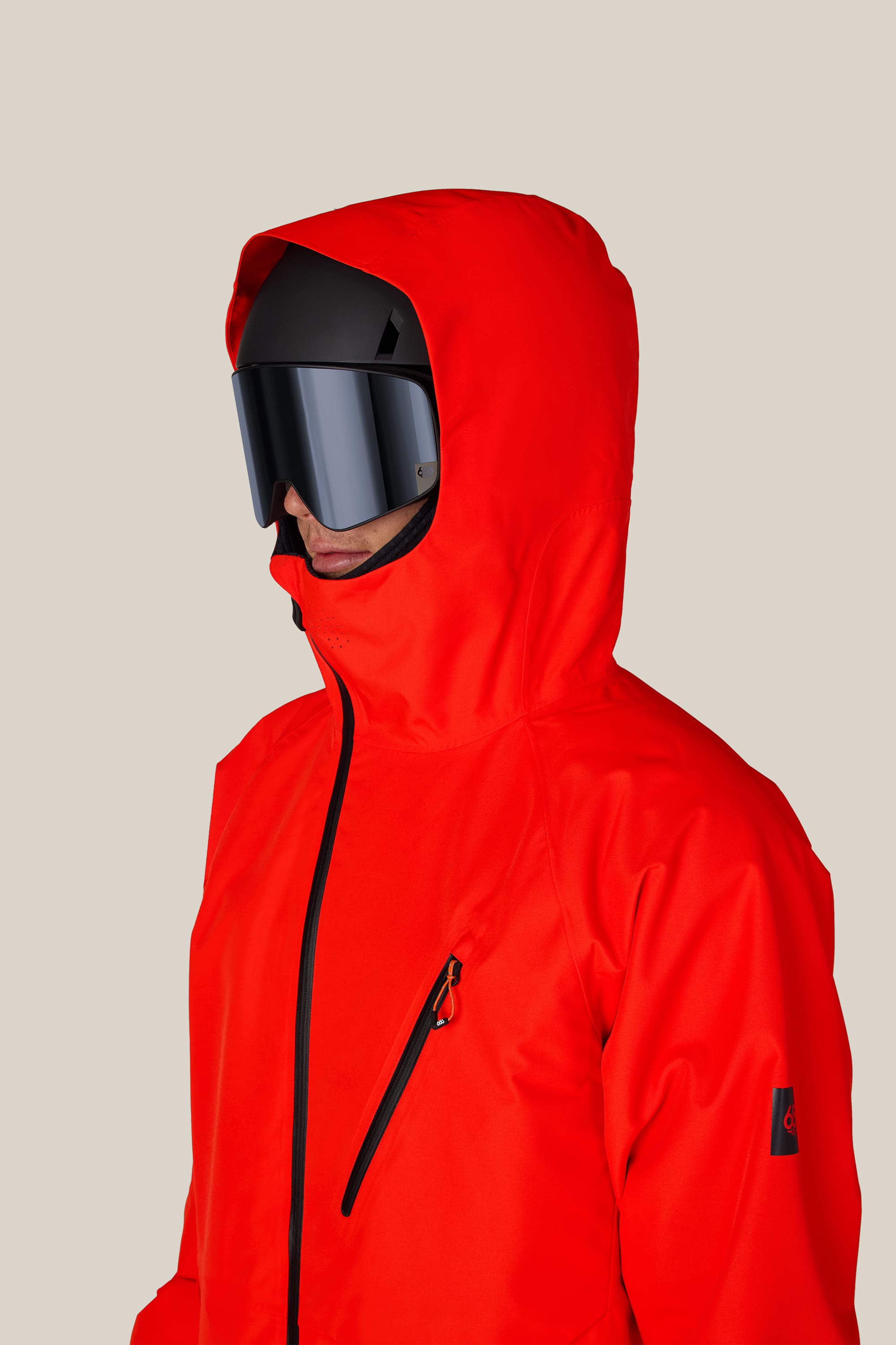 A person wearing the 686 Men’s Hydra™ Thermagraph® Jacket in bright red and black ski goggles, facing left against a plain beige background.